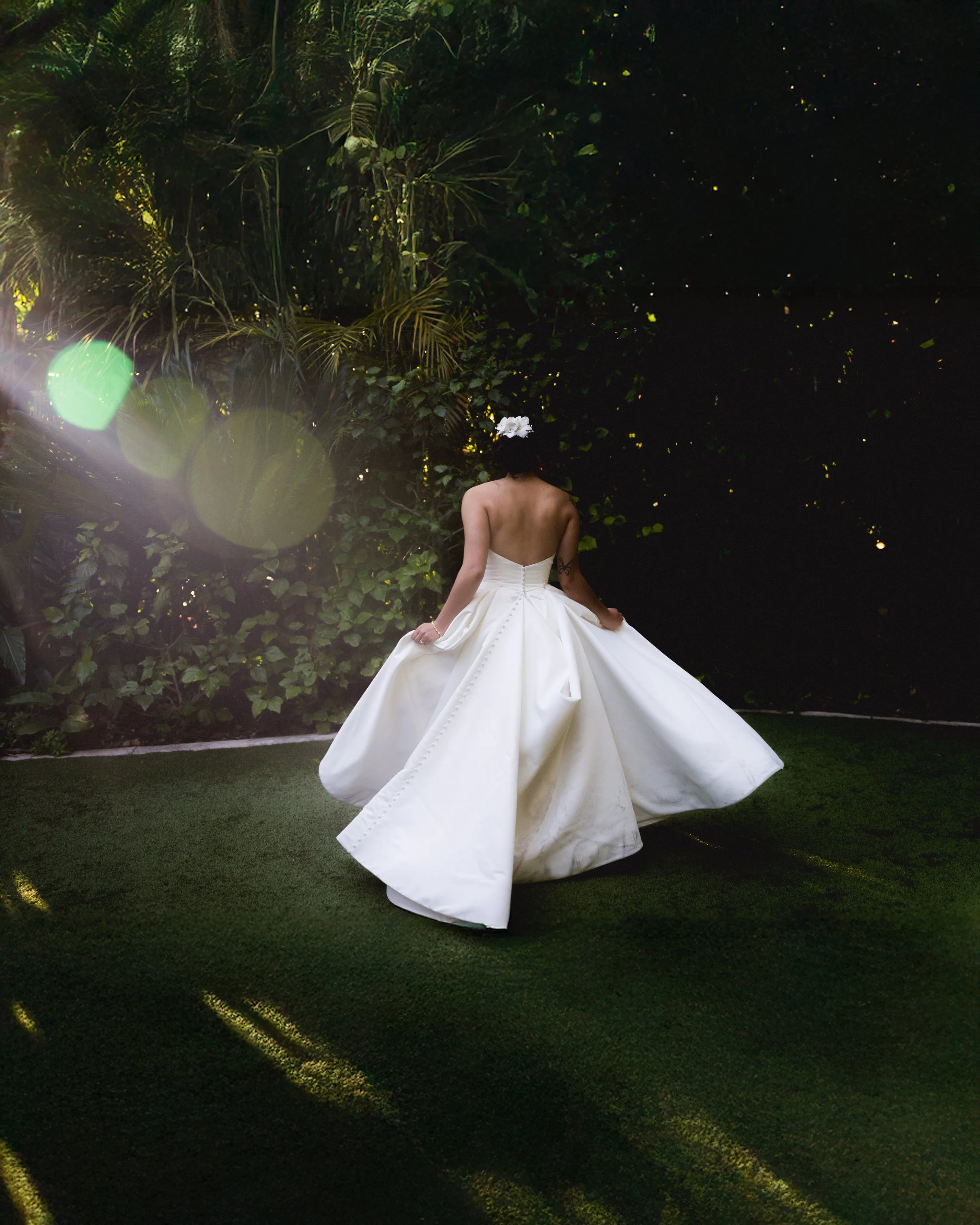 A bride in a strapless white wedding gown with a full skirt, standing outdoors on grass, with her back to the camera. She has a veil or floral headpiece, and her arms are relaxed at her sides. The background features lush green foliage and trees with