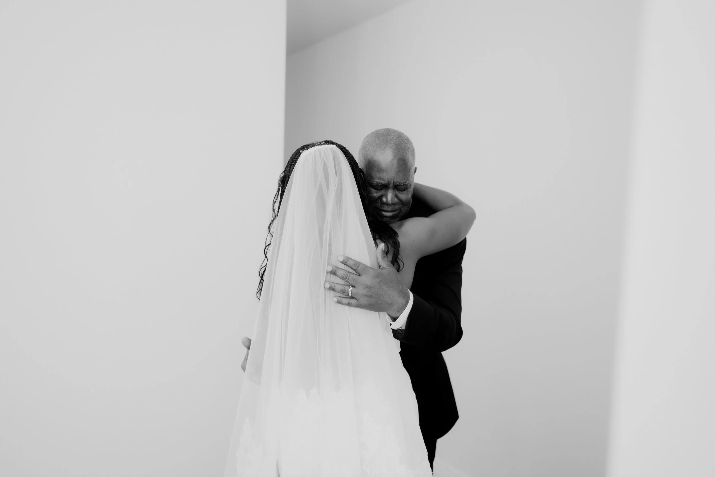 A man in formal attire hugging a woman in a bridal dress and veil, both with emotional expressions, against a plain white background.