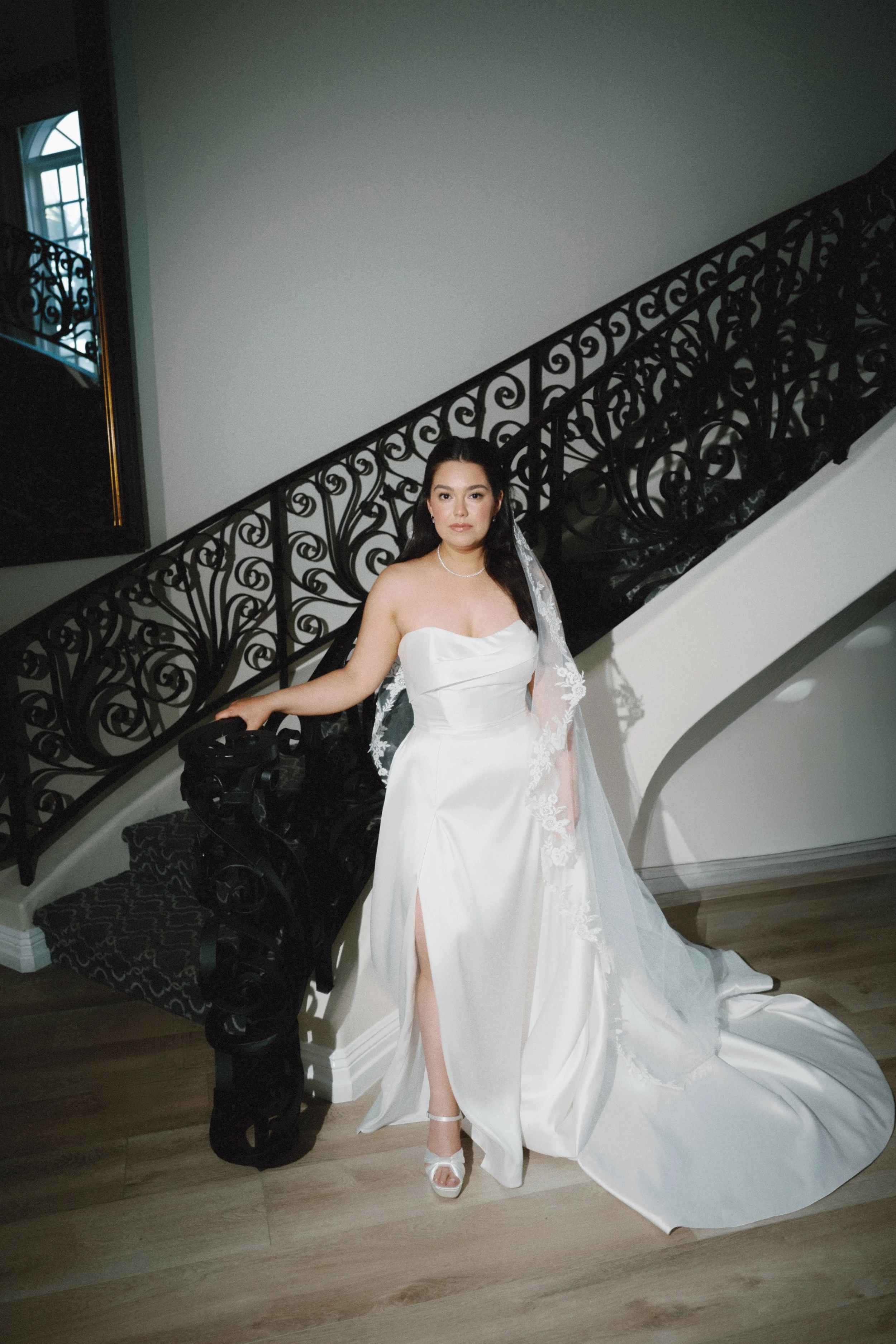 A woman in a white wedding gown standing on a staircase, with black ornate railing, in an elegant interior setting.