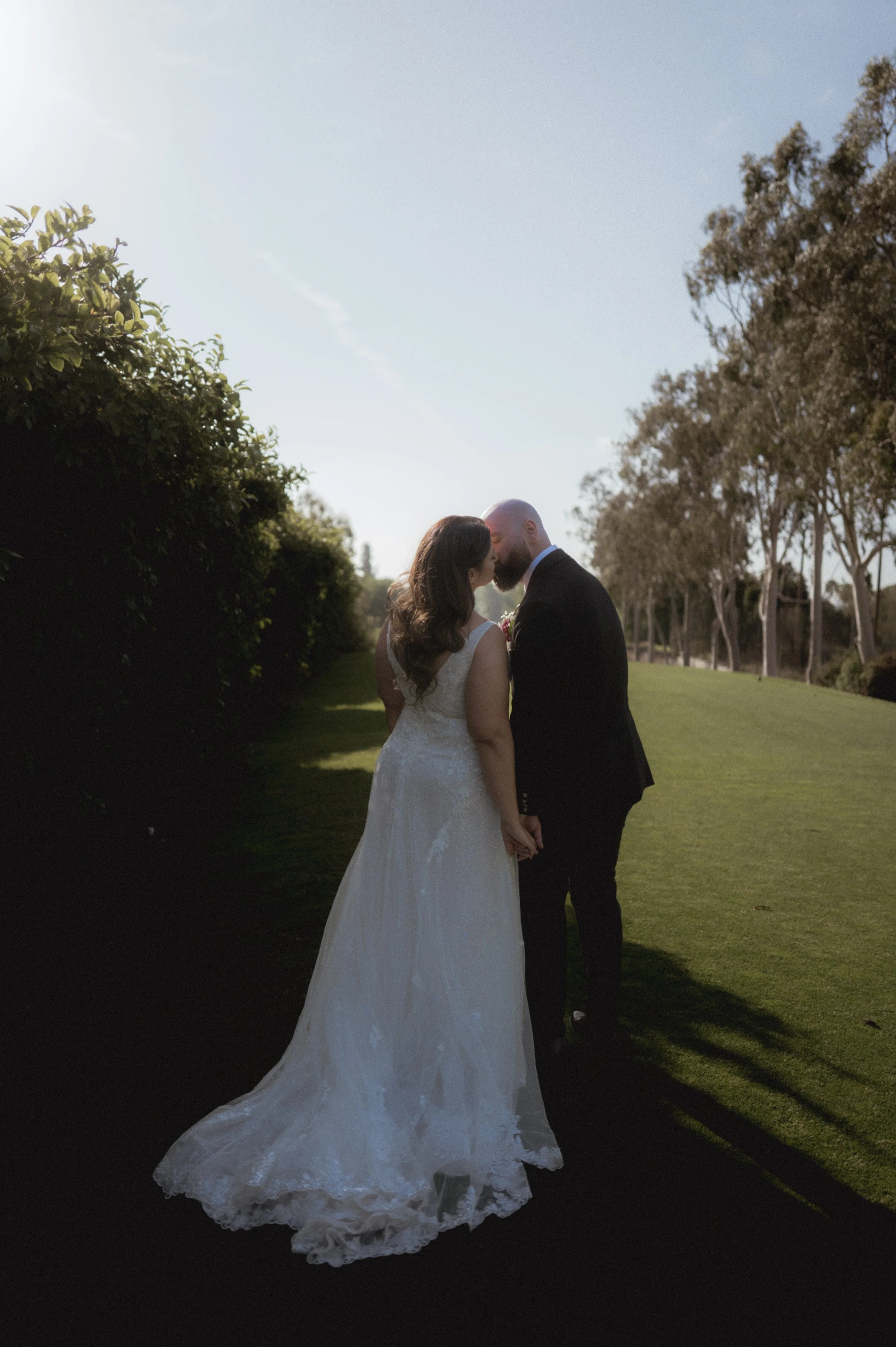 A bride and groom sharing a kiss outdoors on a sunny day, surrounded by trees and green grass.