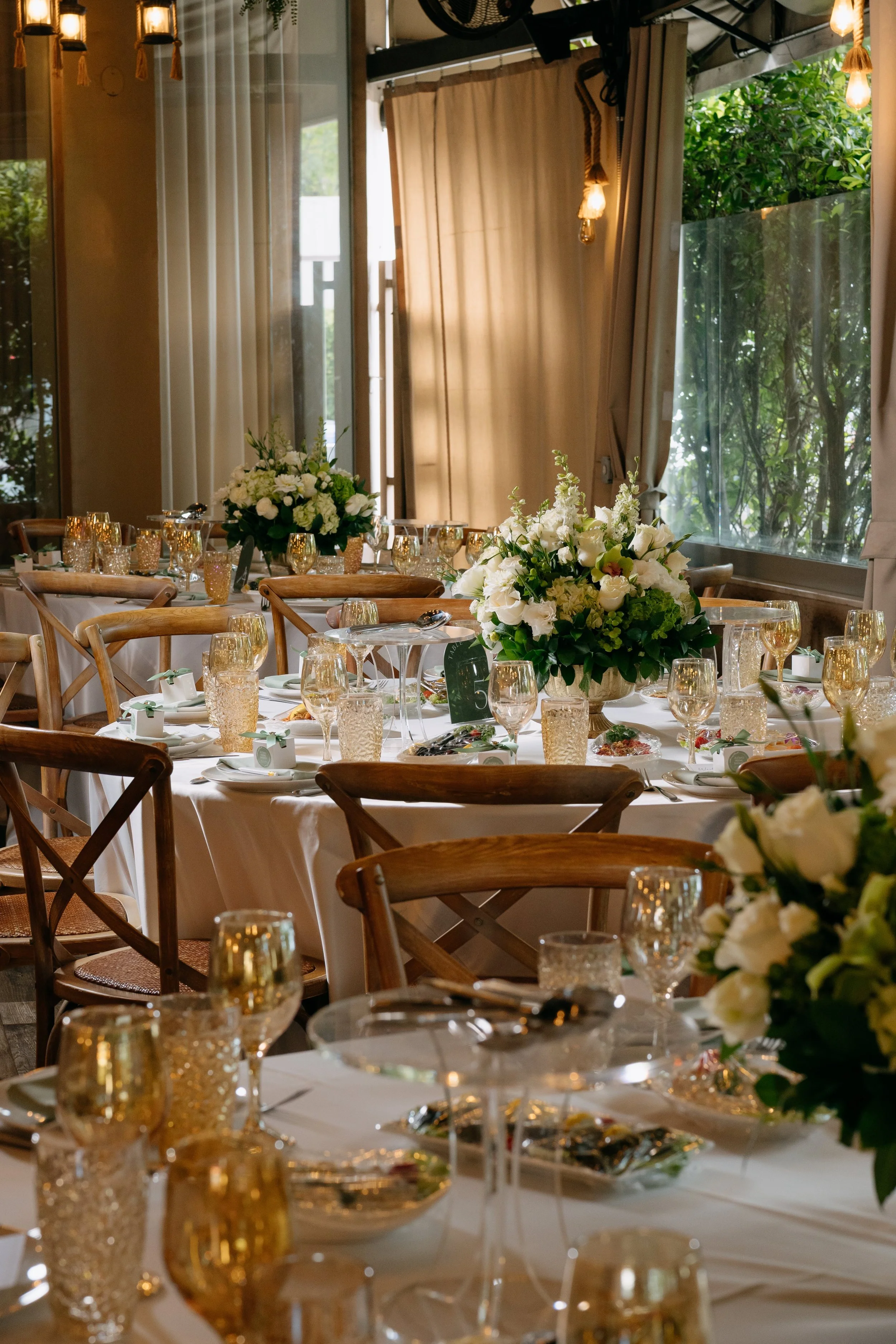 Elegant banquet table setup with white tablecloths, ornate gold-colored glassware, and large floral centerpieces of white flowers and greenery. The setting is indoors with natural light coming through large windows, beige curtains, and warm hanging l