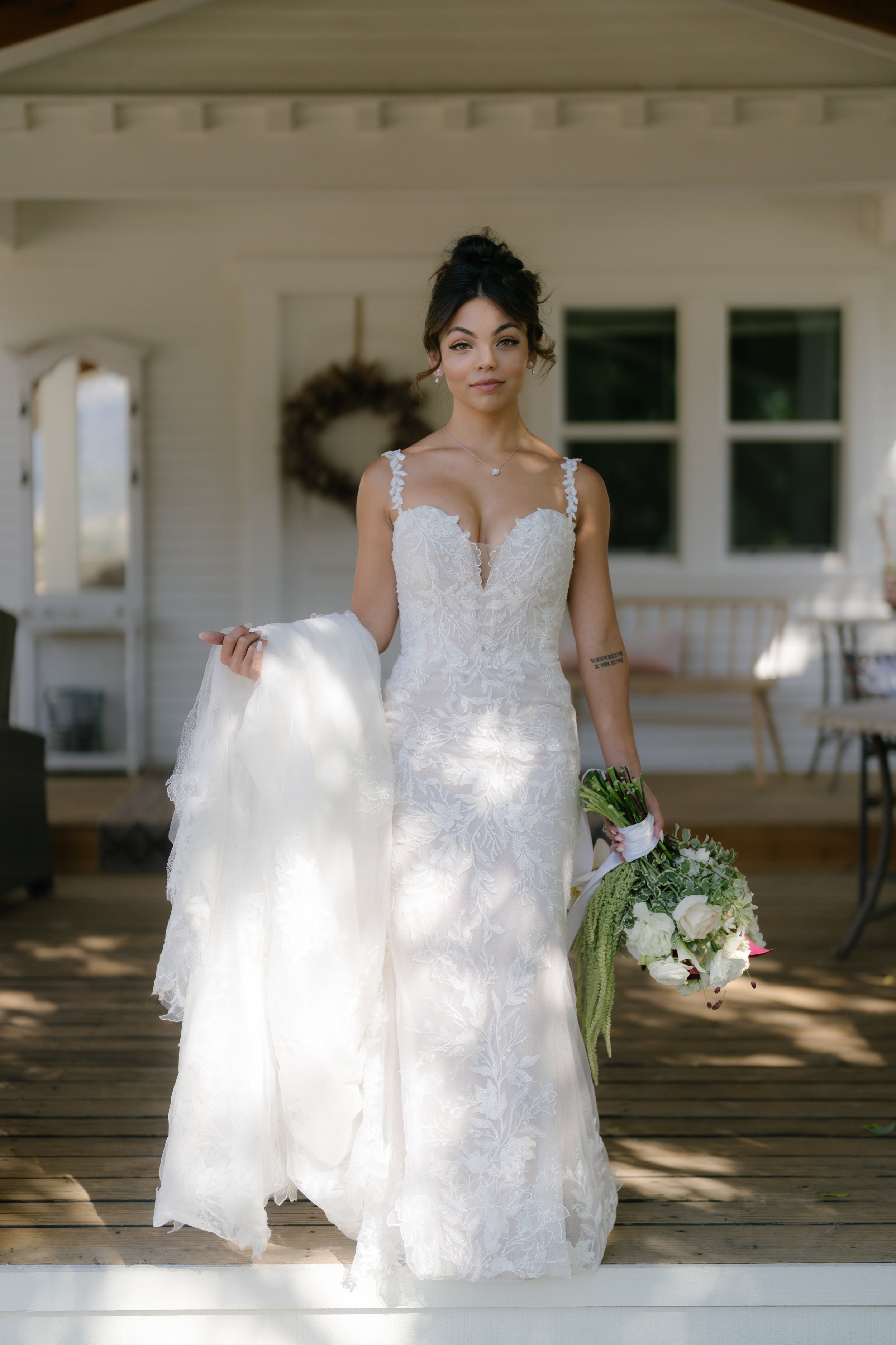 A bride in a white lace wedding dress holding a bouquet of white and green flowers, standing on a wooden porch with white walls and windows behind her.