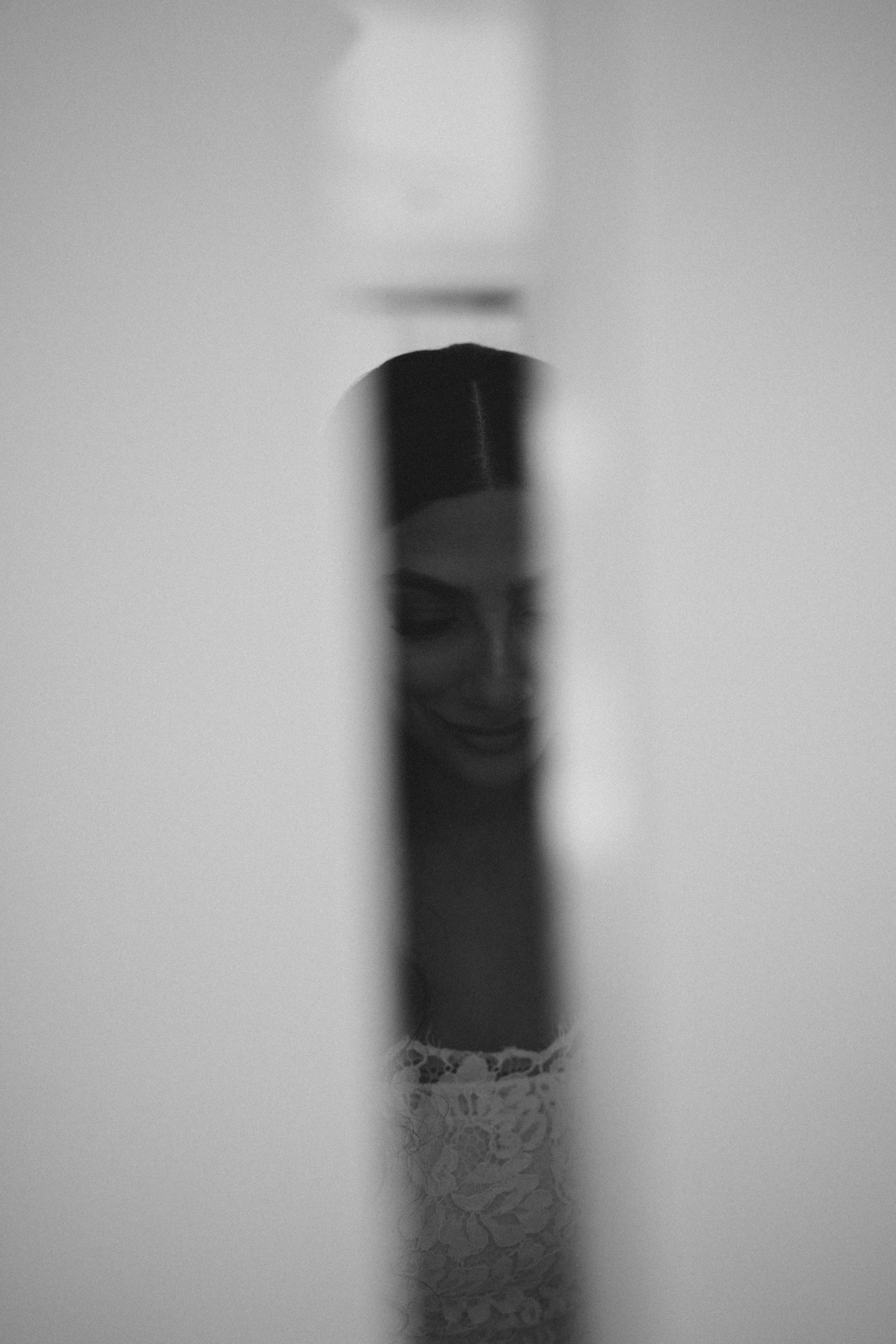 A black and white photo of a woman wearing lace lingerie, with her face partially obscured by her hair, standing in a minimalistic setting with plain walls.