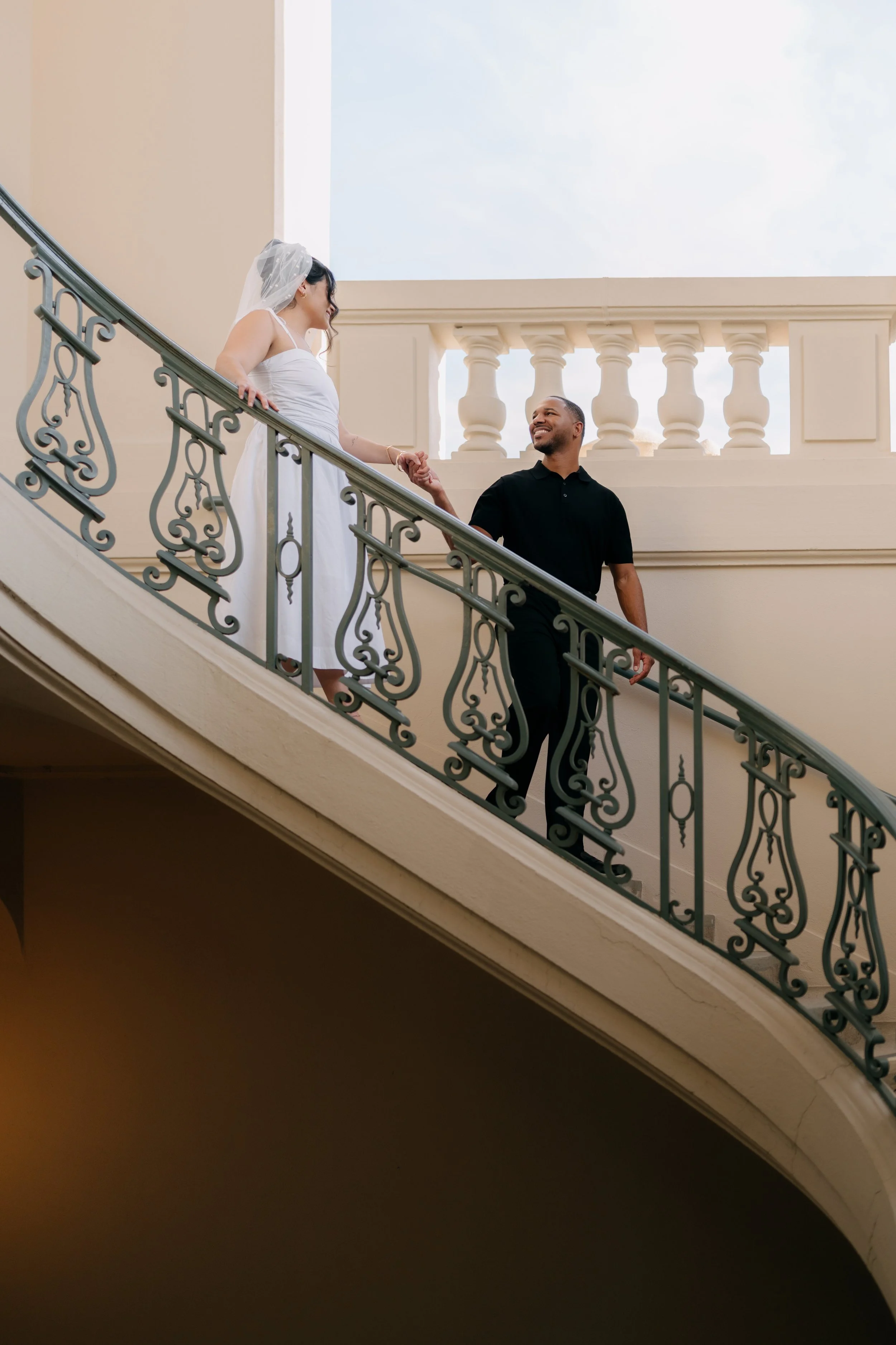 A woman wearing a white wedding dress and veil and a man wearing a black shirt, holding hands, standing on a staircase with decorative railing, in an elegant architectural setting with balustrades and large windows.