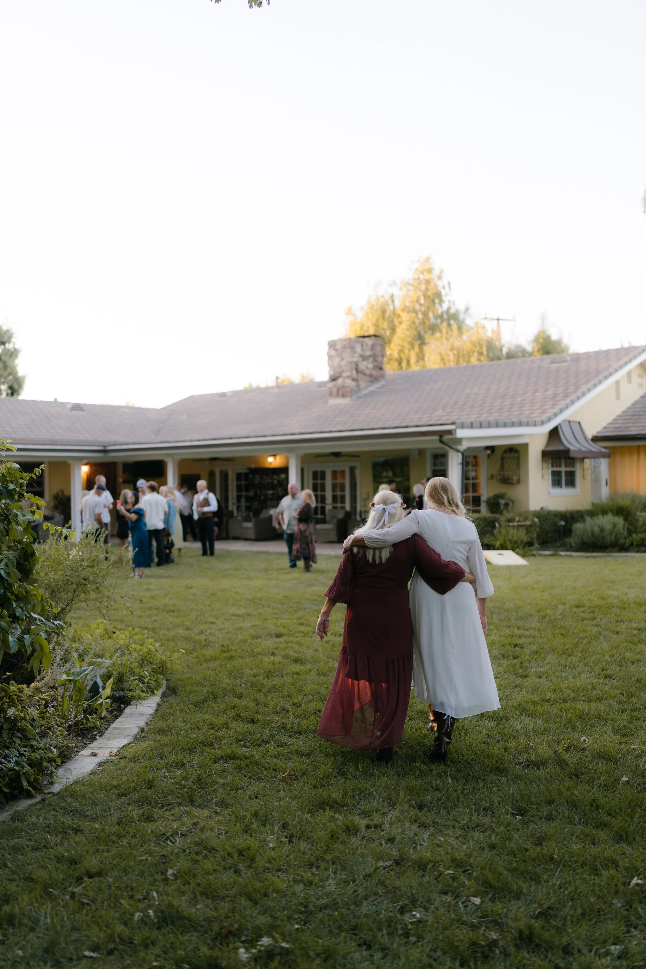 Two women walk arm in arm on a lawn in front of a house during daylight. One woman has long blonde hair and is dressed in a white dress, and the other has white or gray hair and is dressed in a burgundy dress. There are people gathered and chatting i