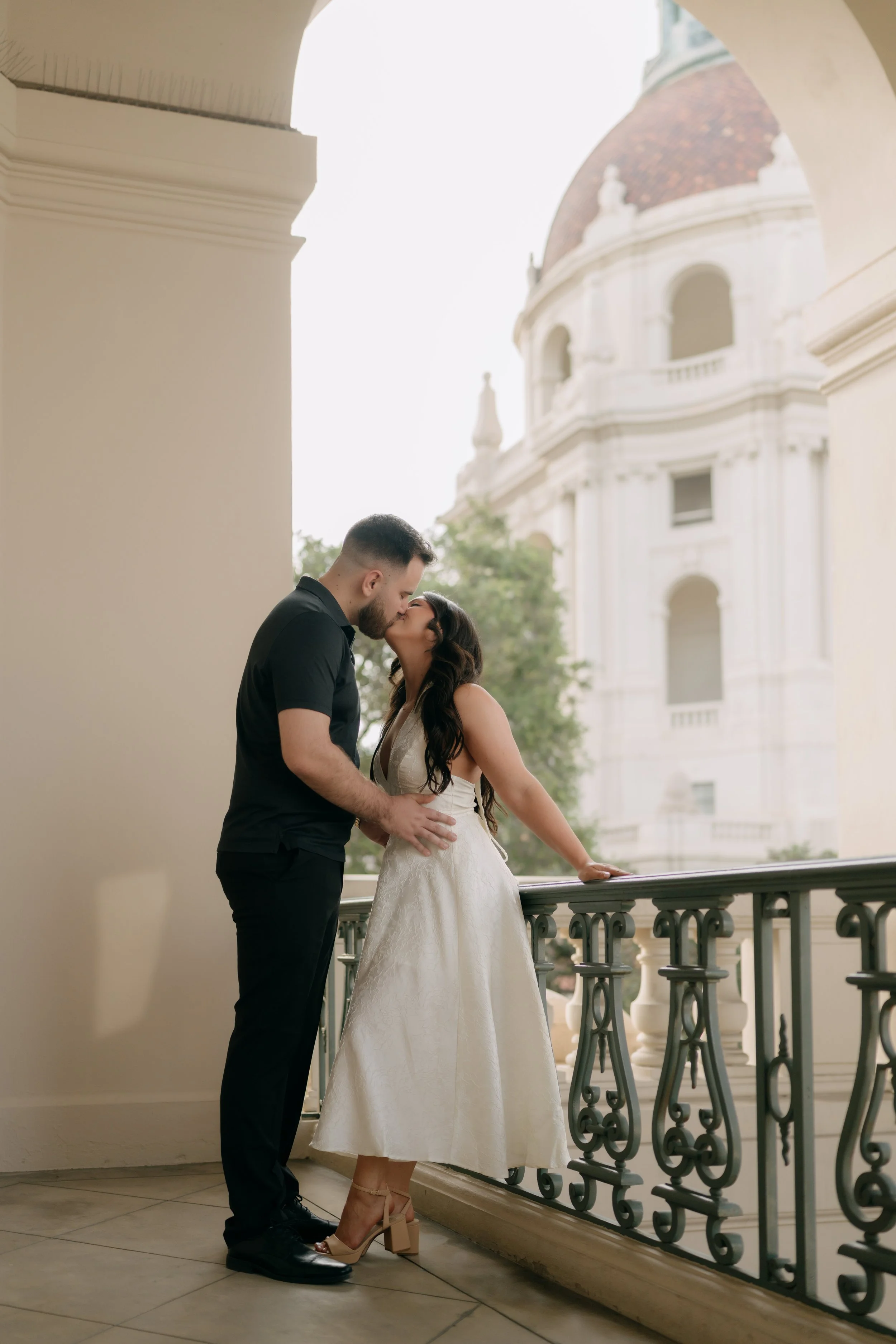 A man and woman sharing a kiss on a balcony overlooking a historic building with a domed roof.