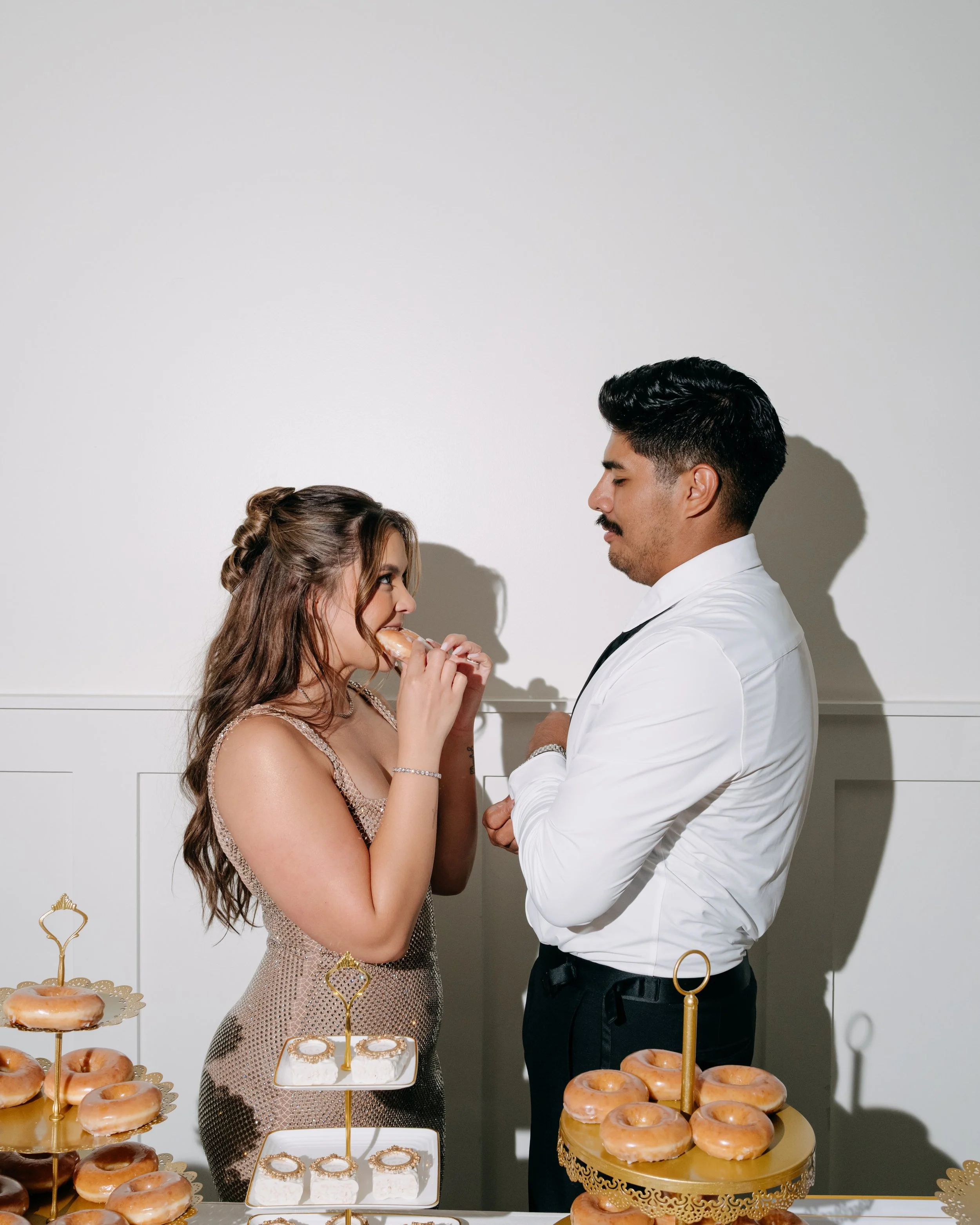 A young woman and a man are standing face to face, gazing into each other's eyes, at a dessert table with donuts and decorated treats.