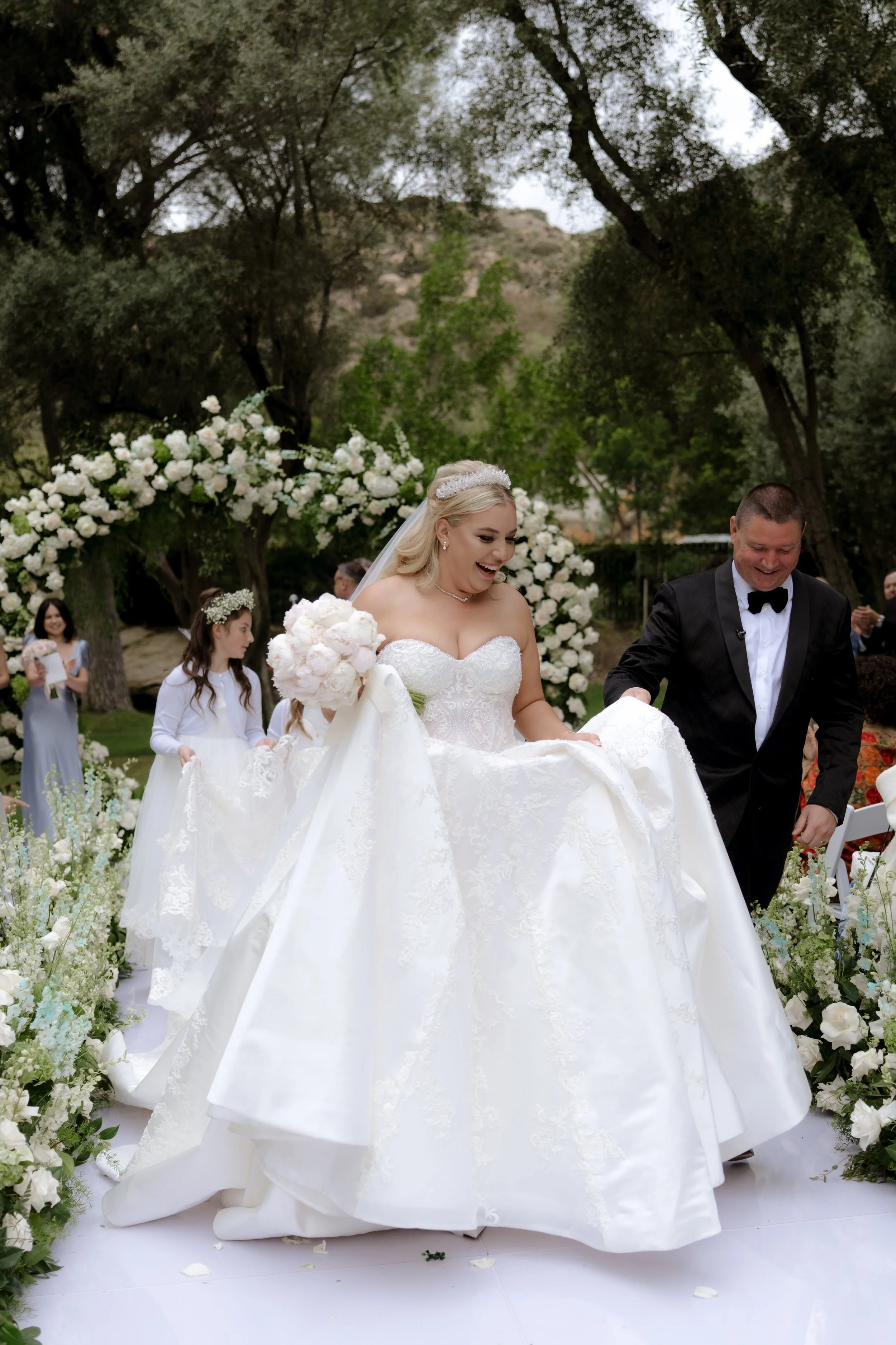 A bride in a white wedding gown with a veil and tiara, holding a bouquet, smiling as she walks down the aisle at her outdoor wedding ceremony. The groom, dressed in a black tuxedo, stands beside her.