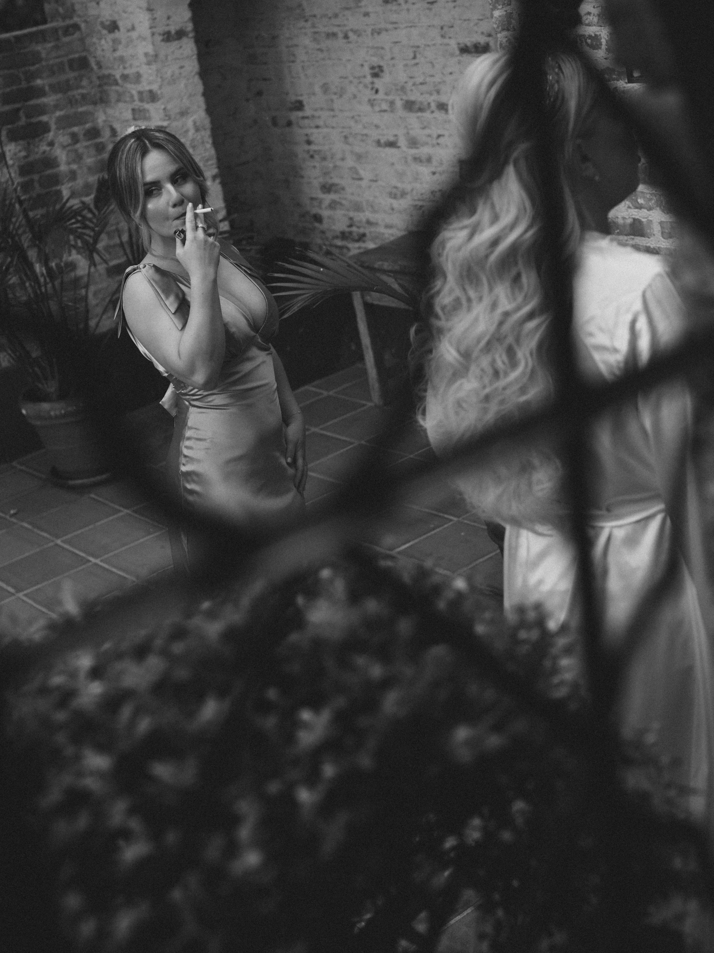 A woman with wavy hair and wearing a silky dress sitting against a brick wall, smoking a cigarette, with plants nearby, in a dimly lit setting.