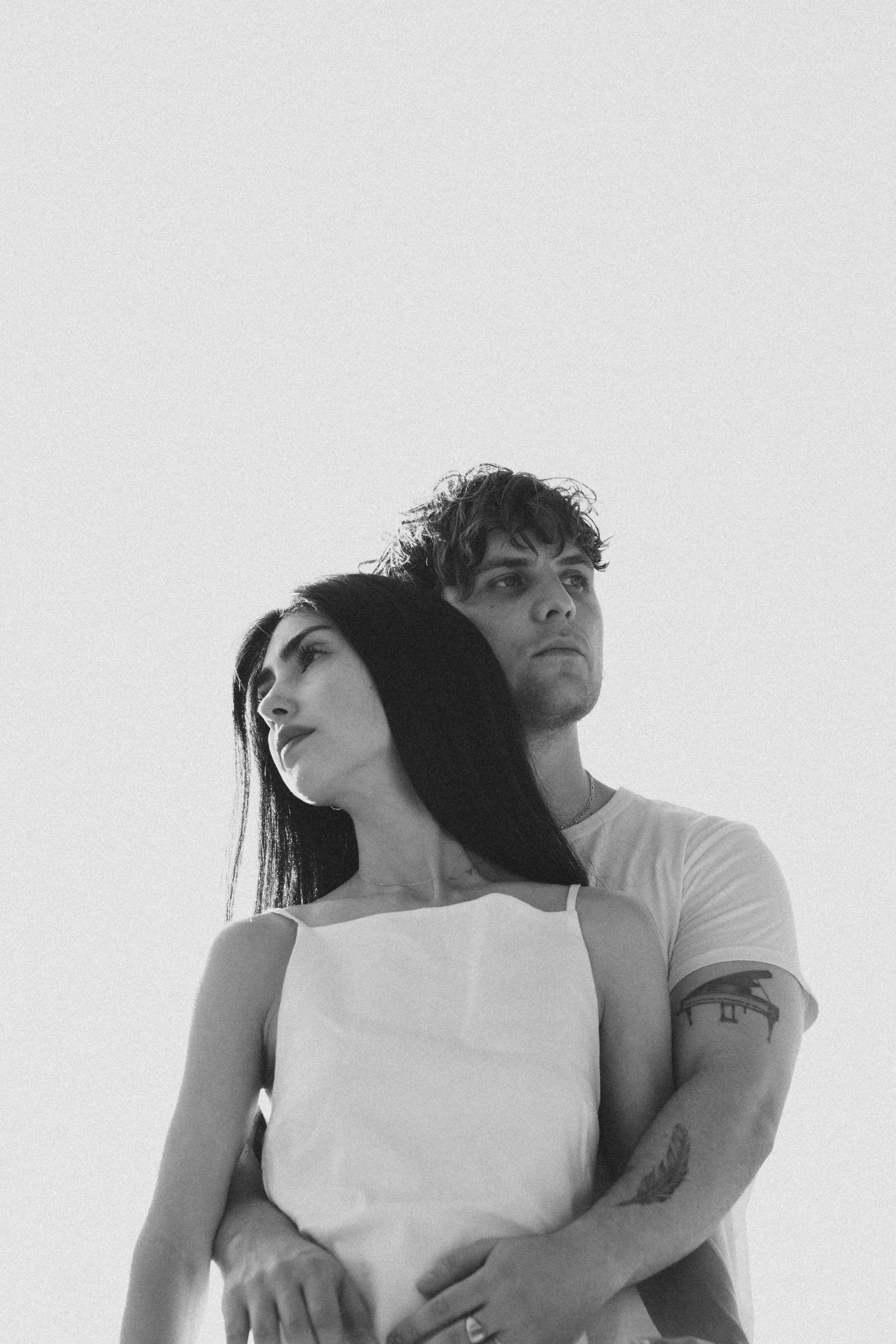 A black-and-white photo of a young woman with long dark hair and a young man with curly hair, both looking to the side. The woman has her arms crossed, and the man is standing behind her with his hand on her shoulder. The woman has a tattoo on her ar