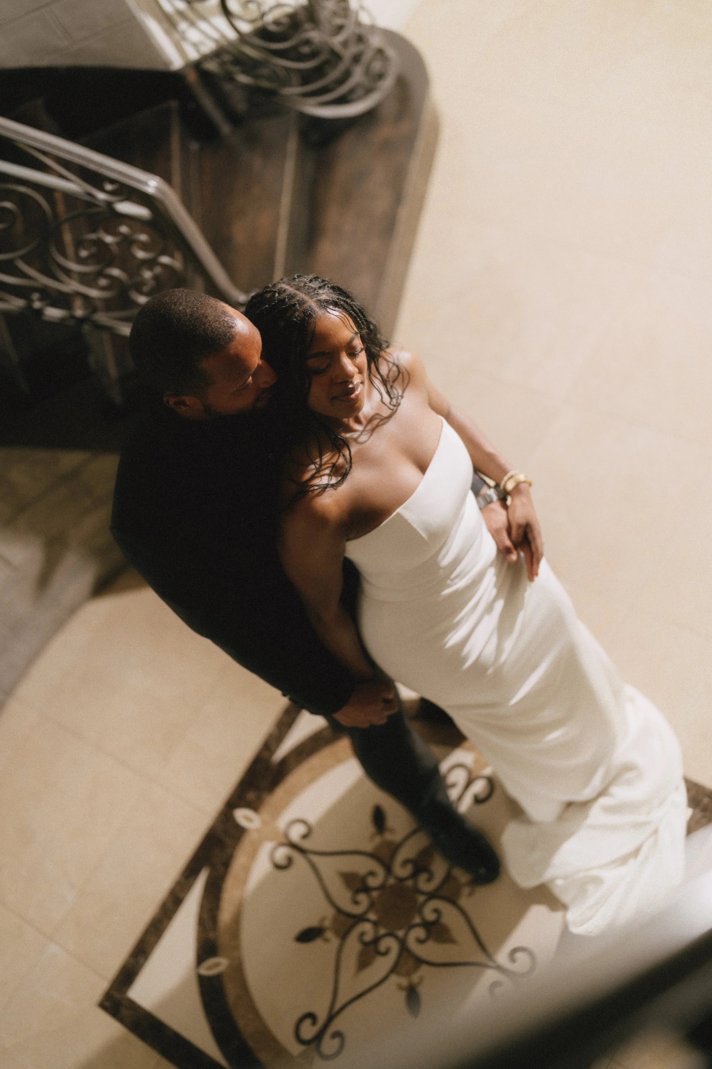 A bride and groom embrace, standing on a decorated staircase, with the bride in a strapless white dress and the groom in a dark suit.