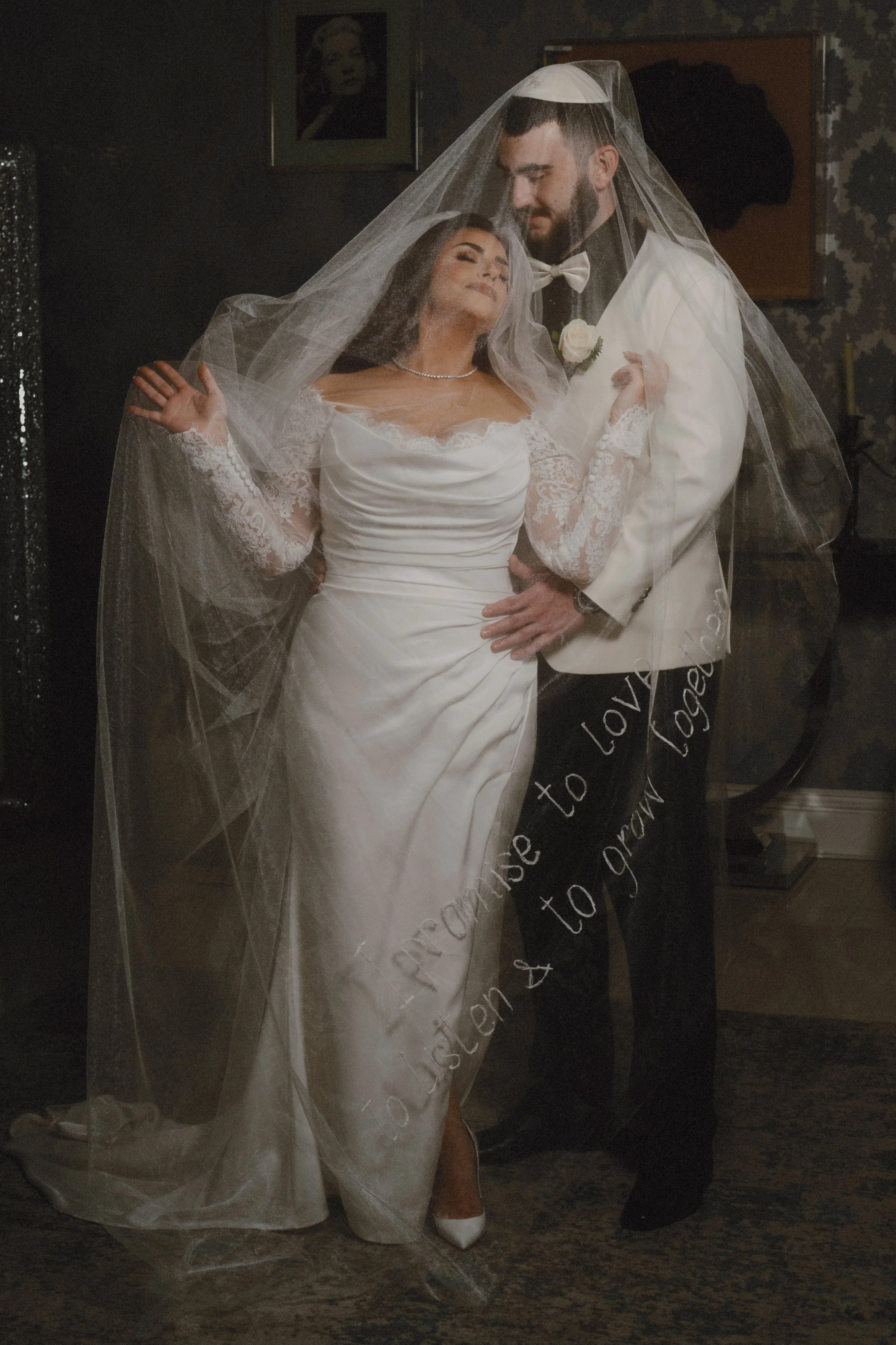 A bride and groom sharing a tender moment during their wedding, with the bride in a white wedding dress and veil, and the groom in a white tuxedo with a bow tie, both smiling softly.