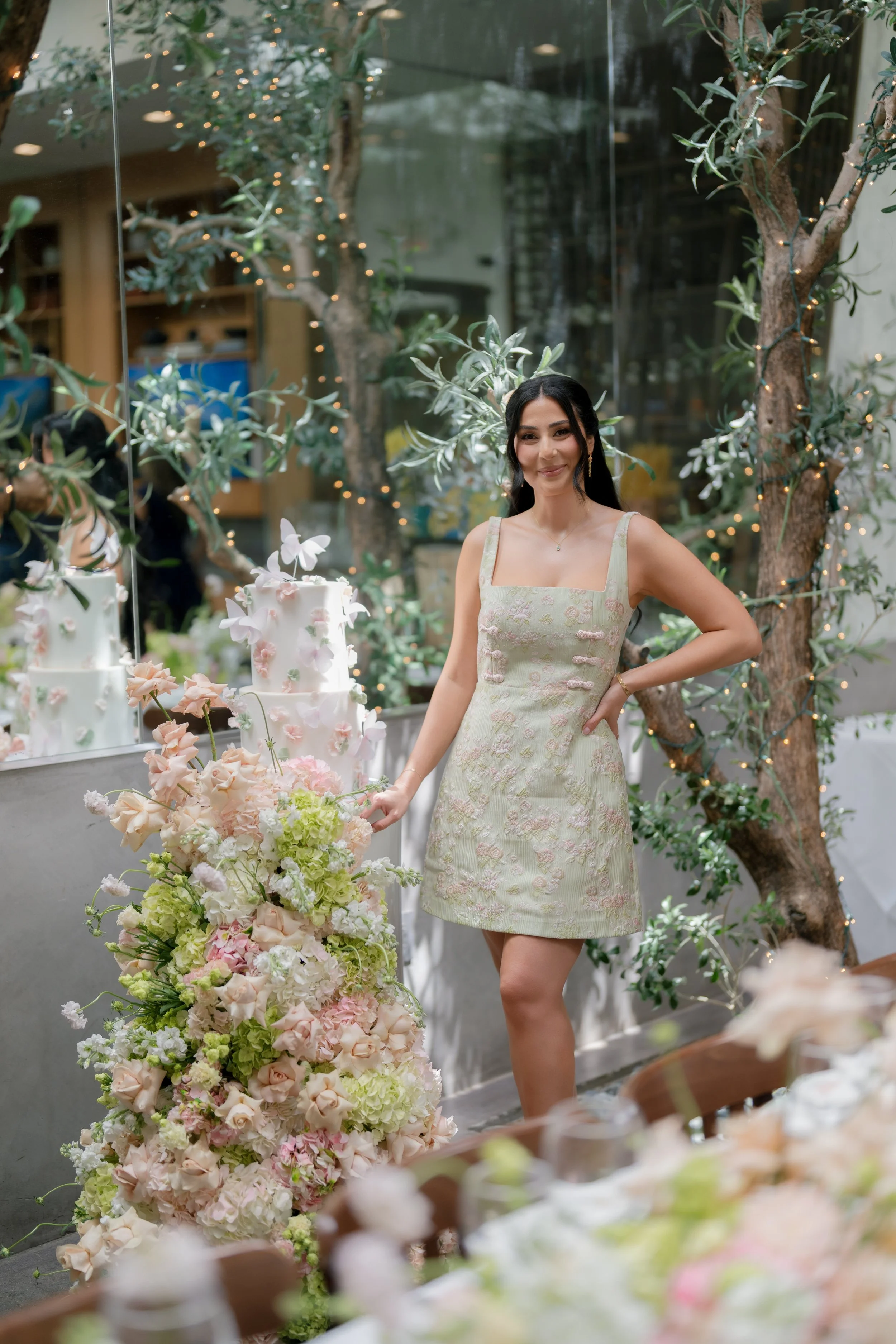 A woman with black hair, wearing a pale green floral dress, standing in front of a display of pastel-colored flowers and cake decorations. She is smiling and surrounded by trees with string lights.
