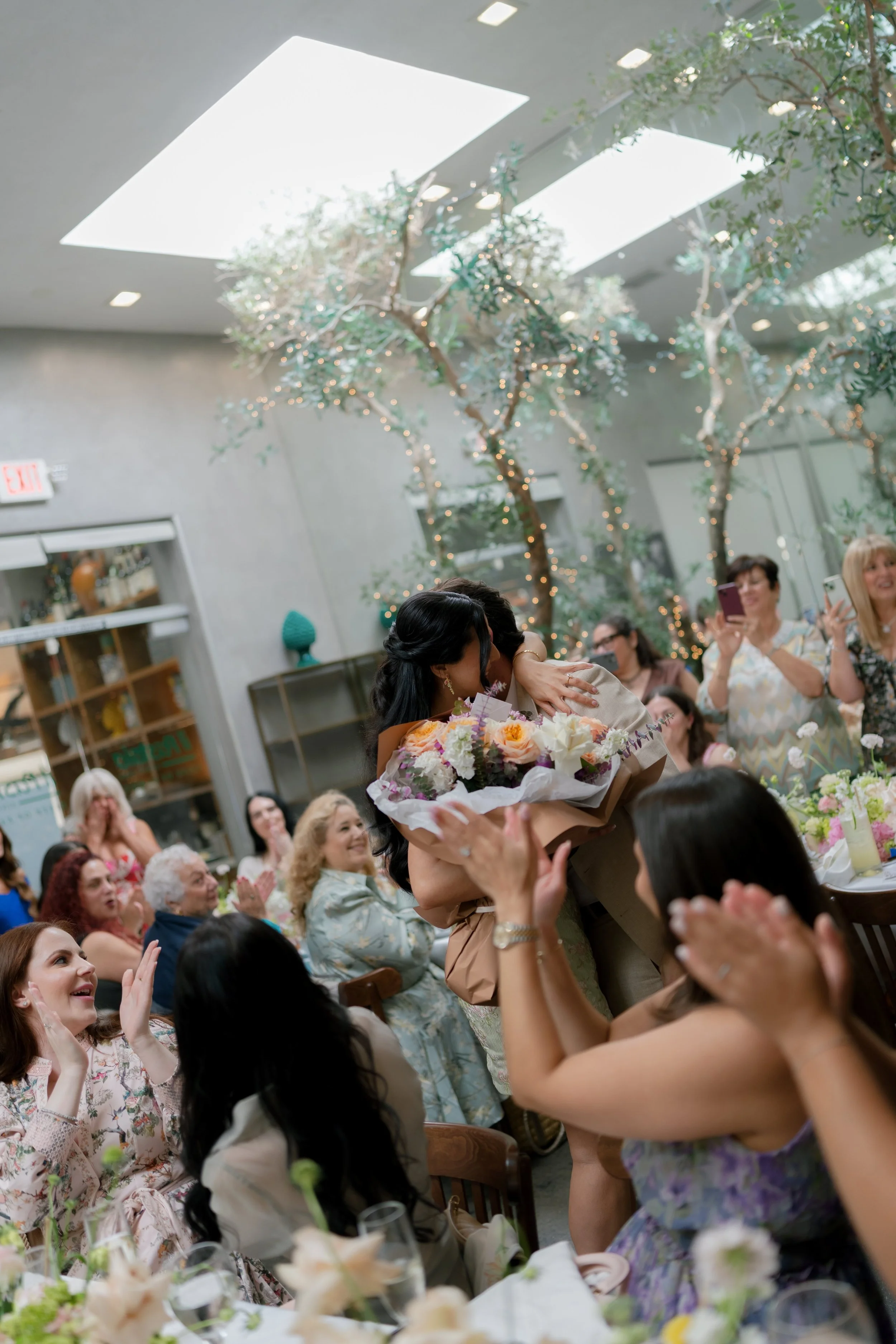 A woman receives a bouquet of flowers at a gathering with seated and standing guests, some taking photos, in a decorated indoor venue with skylights and hanging lights.