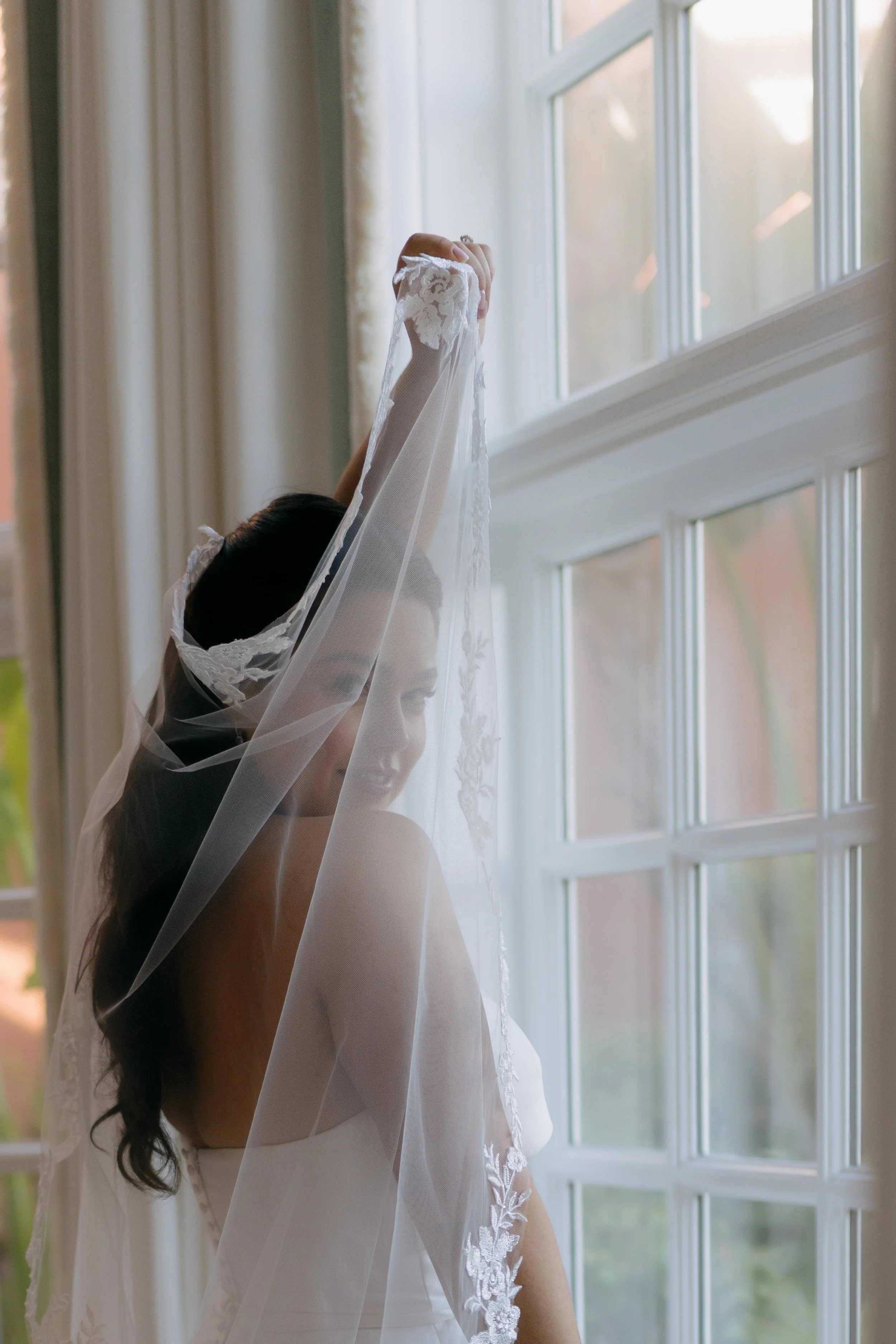 A bride standing by a large window, holding up her lace veil with her right hand, wearing a wedding dress with lace details, with sunlight coming through the window.