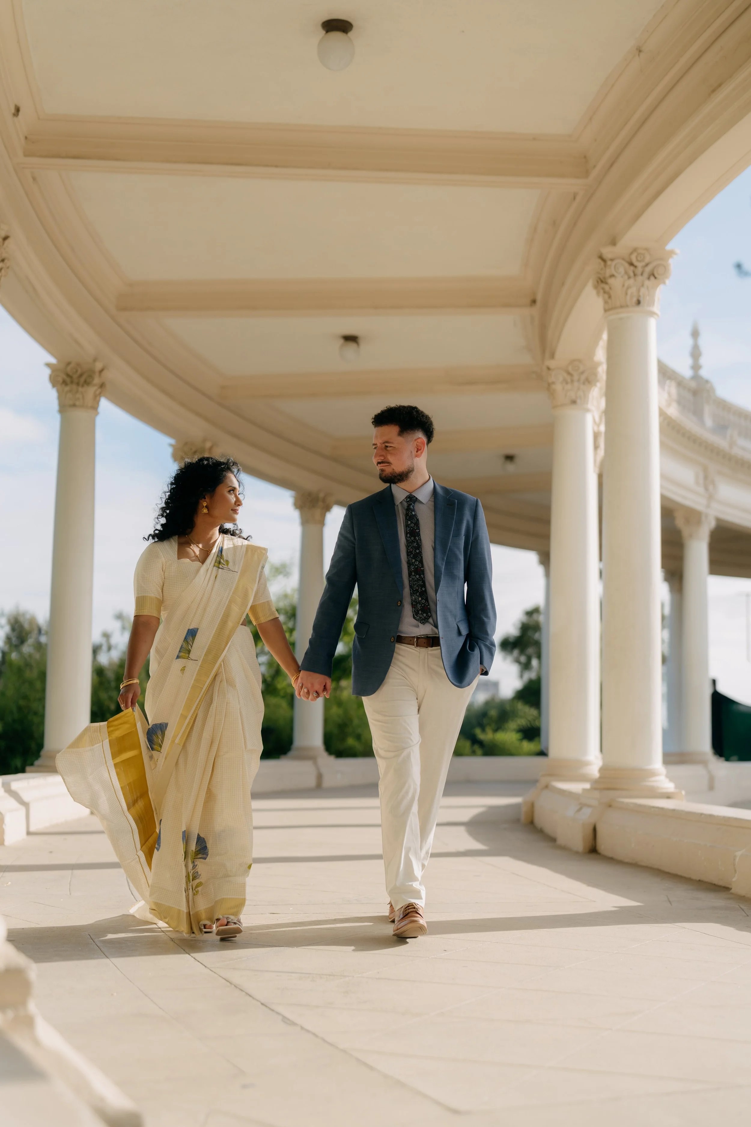 A man and woman holding hands walking under a white classical colonnade with a curved roof, blue sky, and green trees in the background.