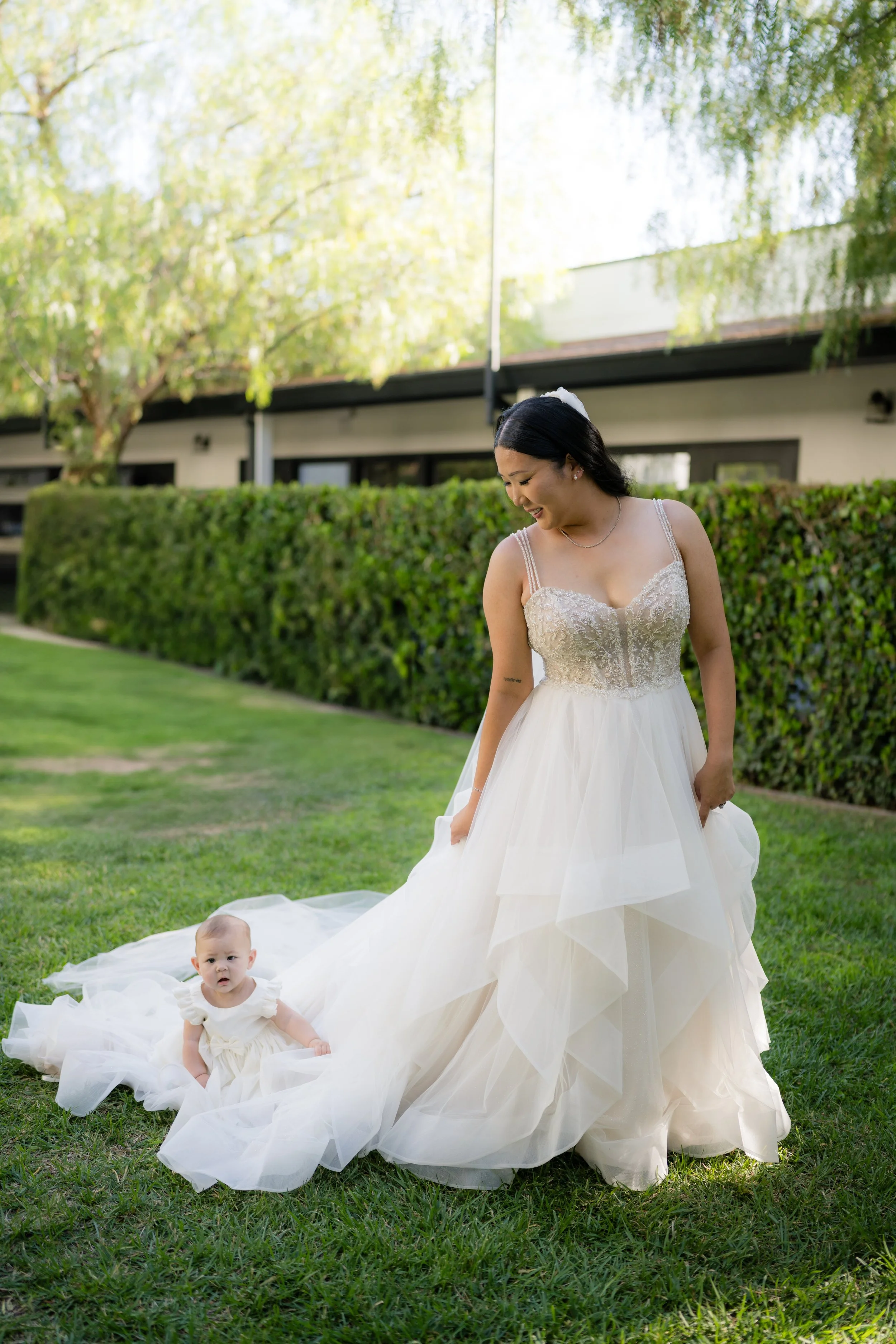 A woman in a wedding dress and a baby girl in a white dress sitting on the grass outdoors, with a green hedge and trees in the background.