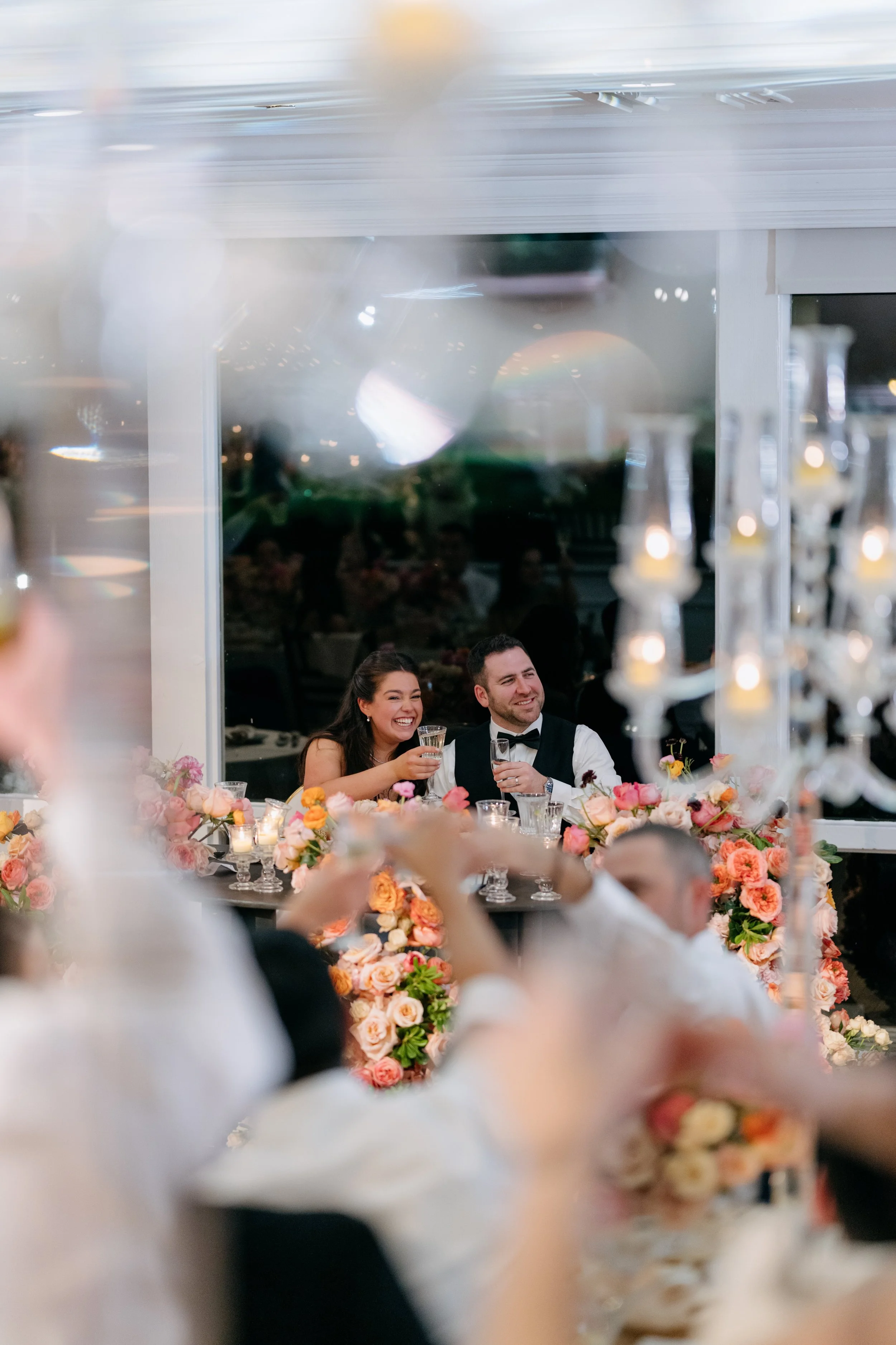 A man and a woman celebrating at a wedding reception, sitting at a table with a floral centerpiece, wearing formal attire, surrounded by guests, with a background of hanging candles and a reflective window.