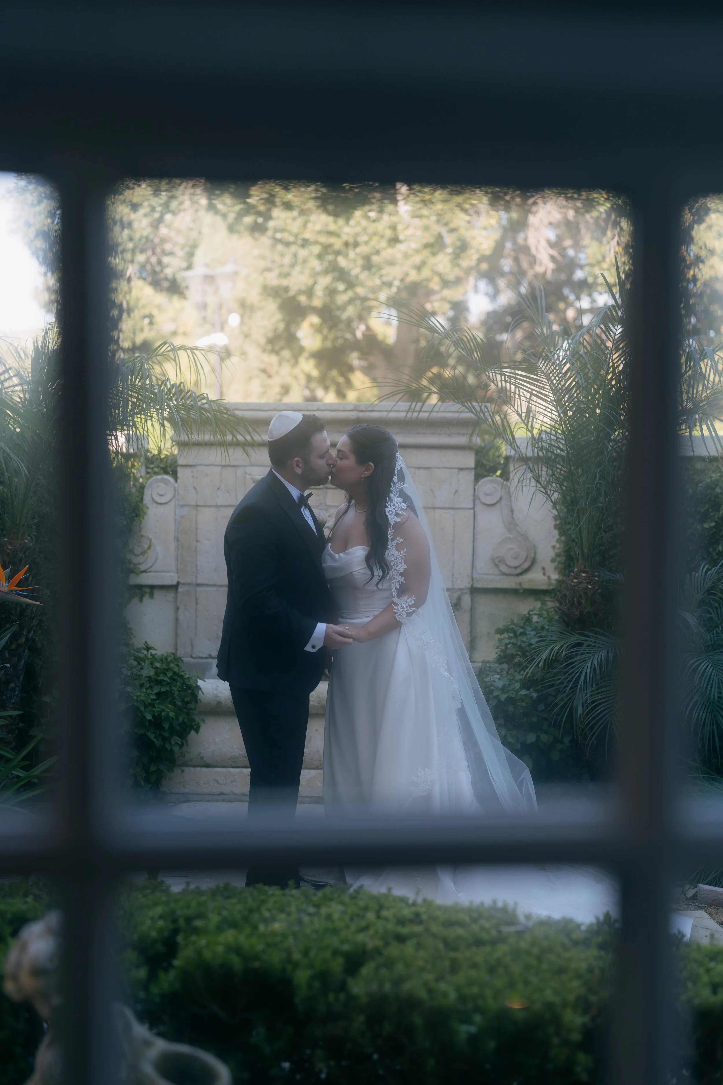 A newlywed couple kissing outdoors in front of trees and lush greenery, viewed through a window or grille.