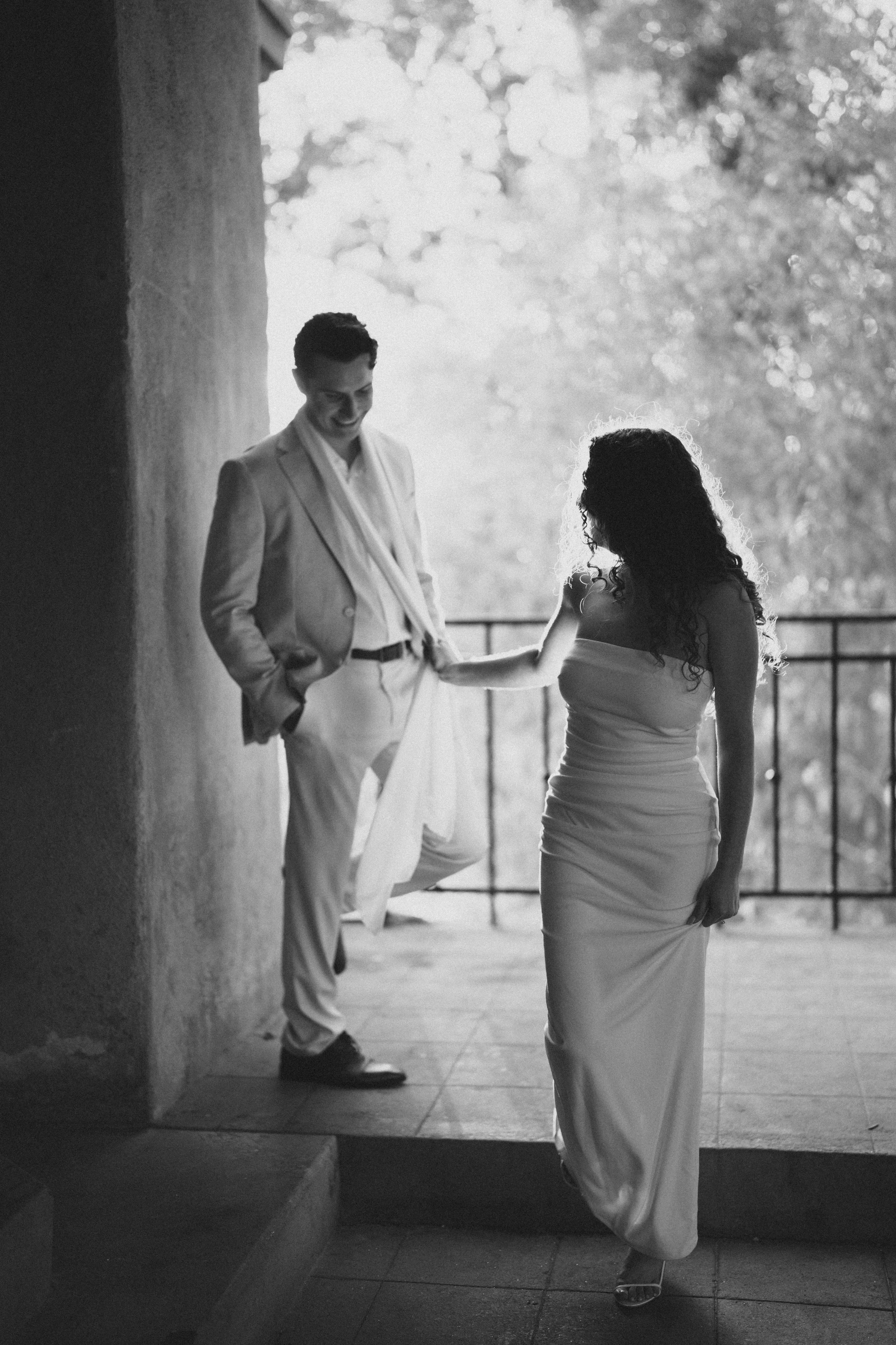A black and white photo of a man and a woman. The man is dressed in a suit and the woman is wearing a strapless dress. They are standing outdoors near a railing, with trees in the background. The man is leaning against a wall, smiling at the woman, w