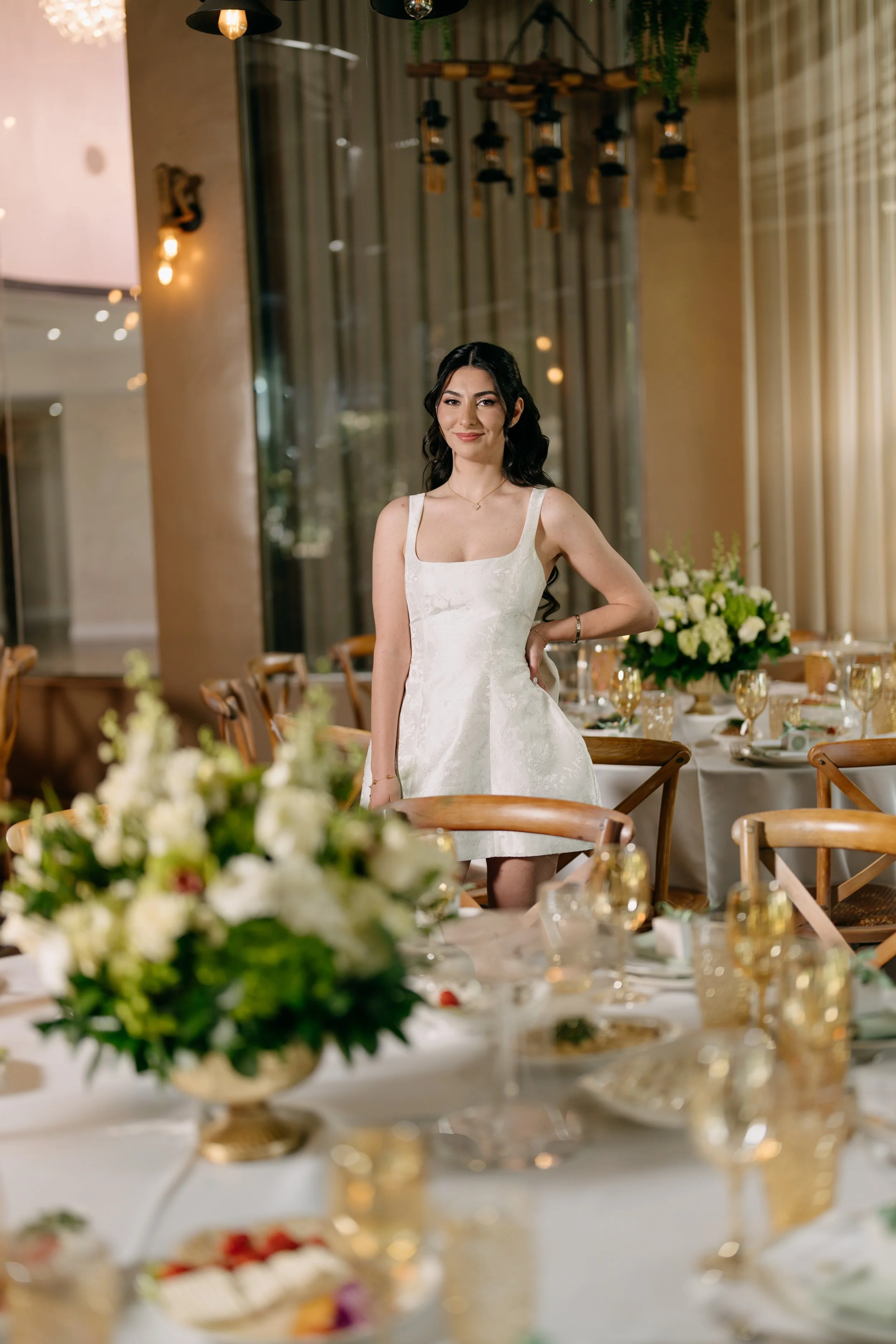 A woman in a white dress stands at a decorated banquet table in a banquet hall, with floral centerpieces and gold-rimmed glassware, during a celebration or wedding event.