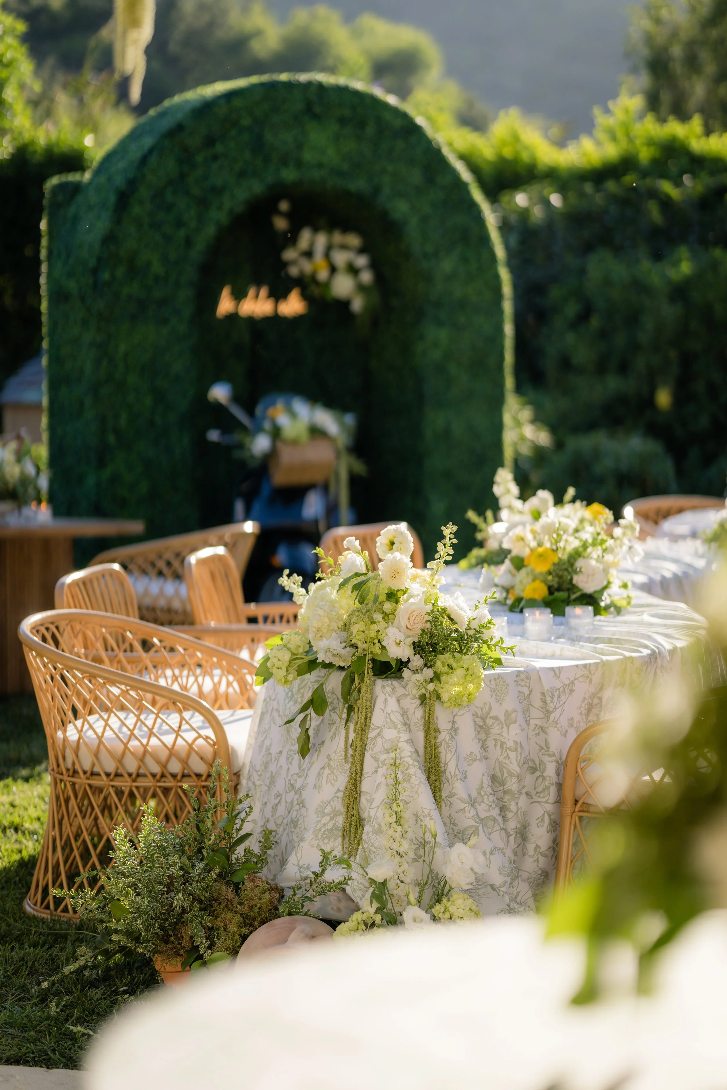 Outdoor wedding reception setup with a round table decorated with white and yellow flowers and greenery, surrounded by wicker chairs. In the background, there is a green hedge arch with flowers and a blurred bicycle, set in a lush garden with trees a