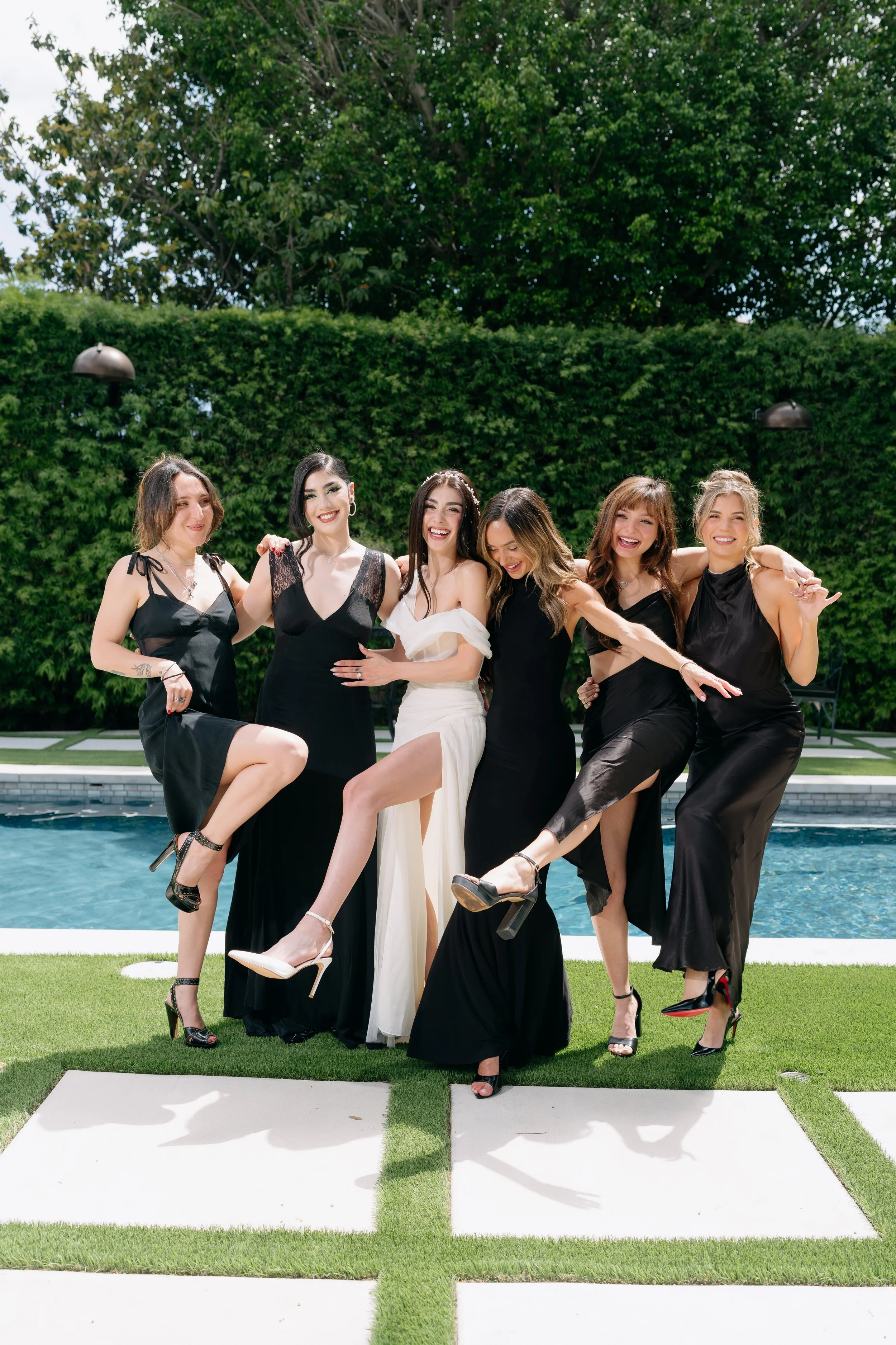 Group of six women dressed in formal black and white dresses posing by a swimming pool on a sunny day, with a background of trees and hedge.