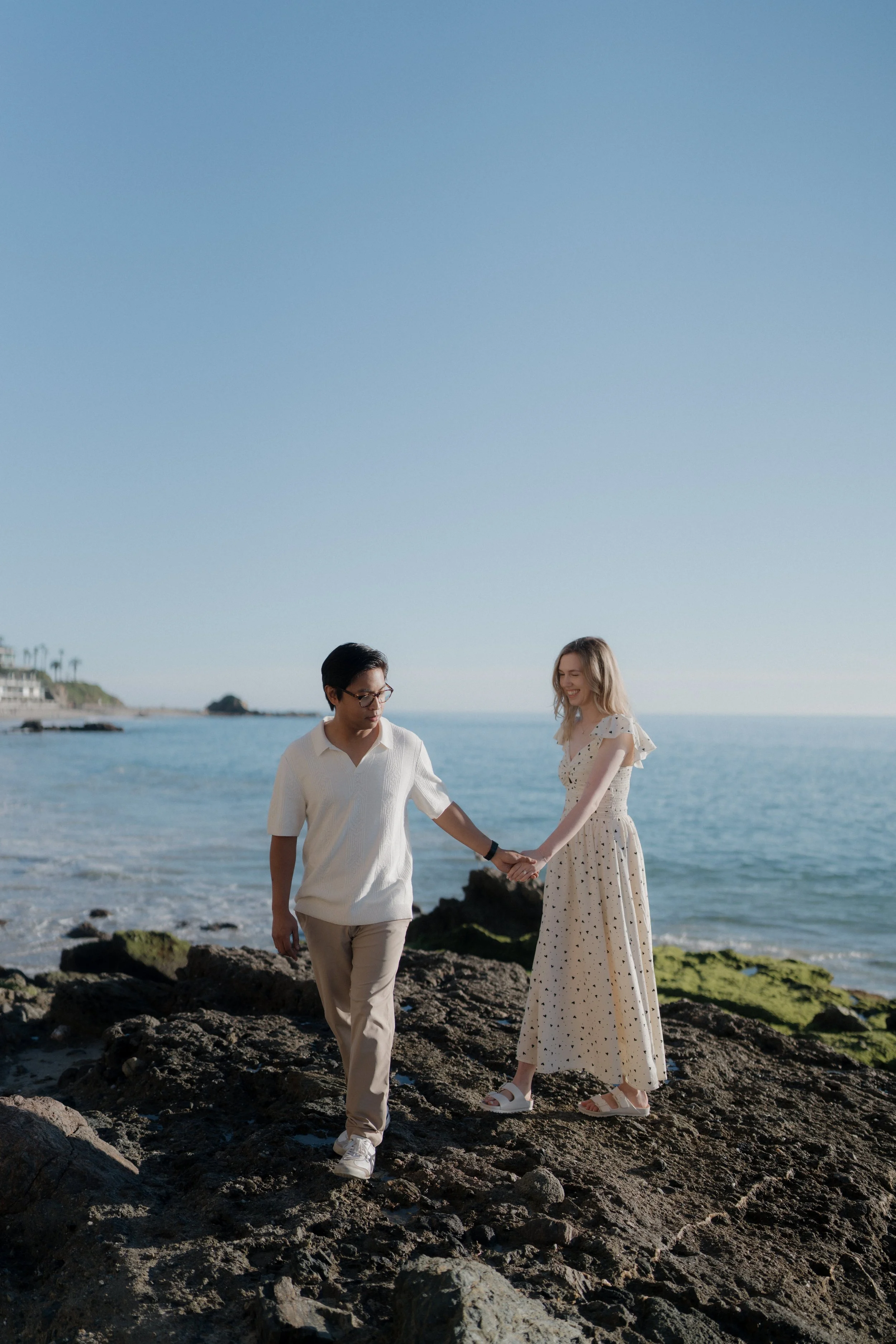 A couple holding hands walking on rocks by the ocean, with a clear blue sky and a distant shoreline with palm trees in the background.