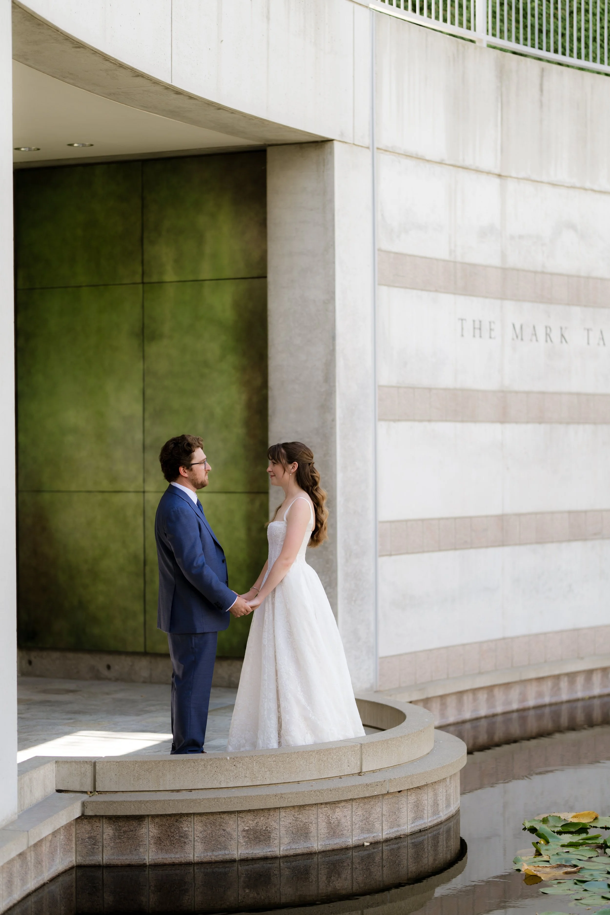 A bride and groom hold hands and face each other on a small circular stone platform near a water fountain with lily pads. The bride wears a white wedding dress, and the groom wears a blue suit with glasses. They stand in front of modern concrete and 