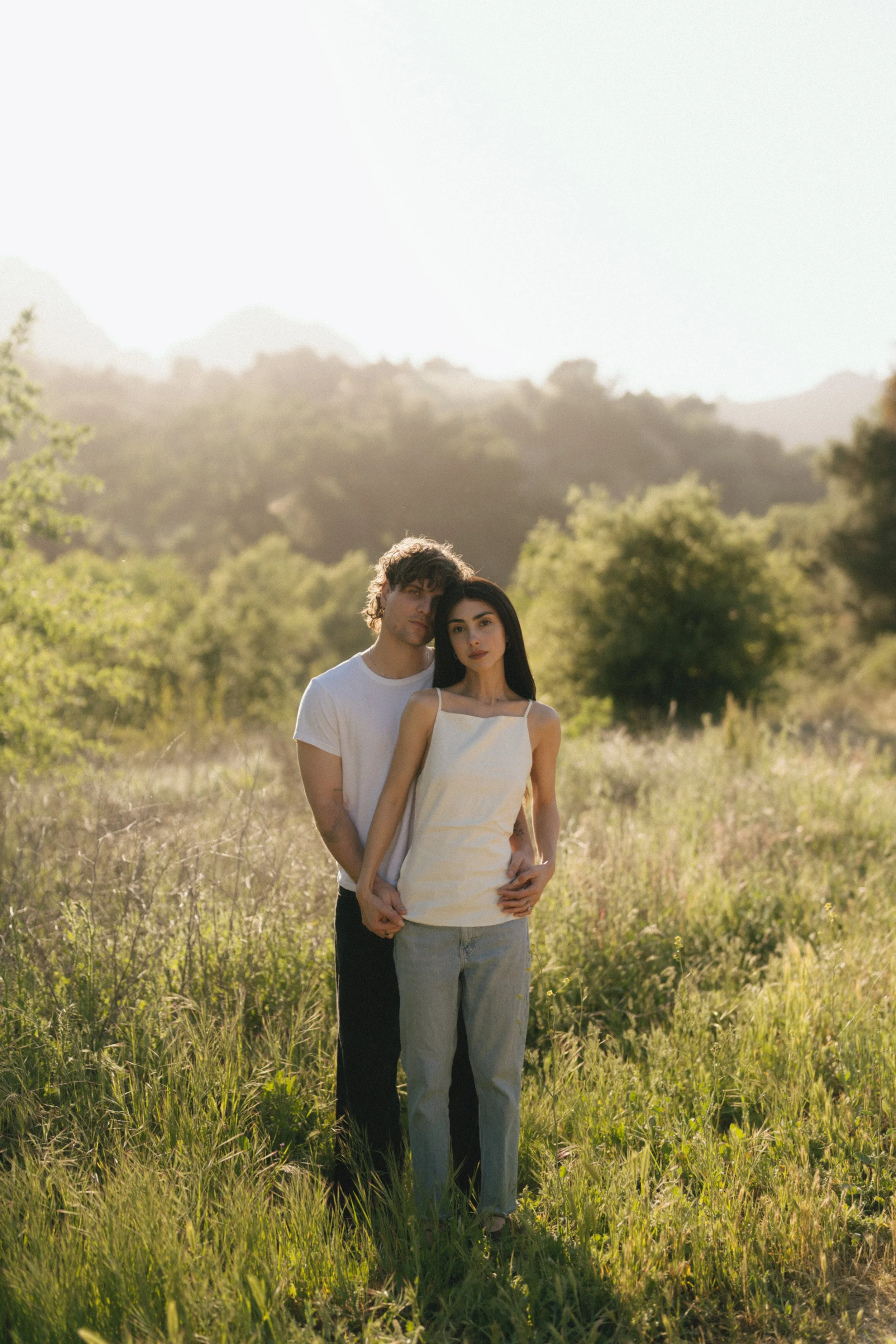 A young couple standing close together in a grassy field with trees and hills in the background, illuminated by warm sunlight.
