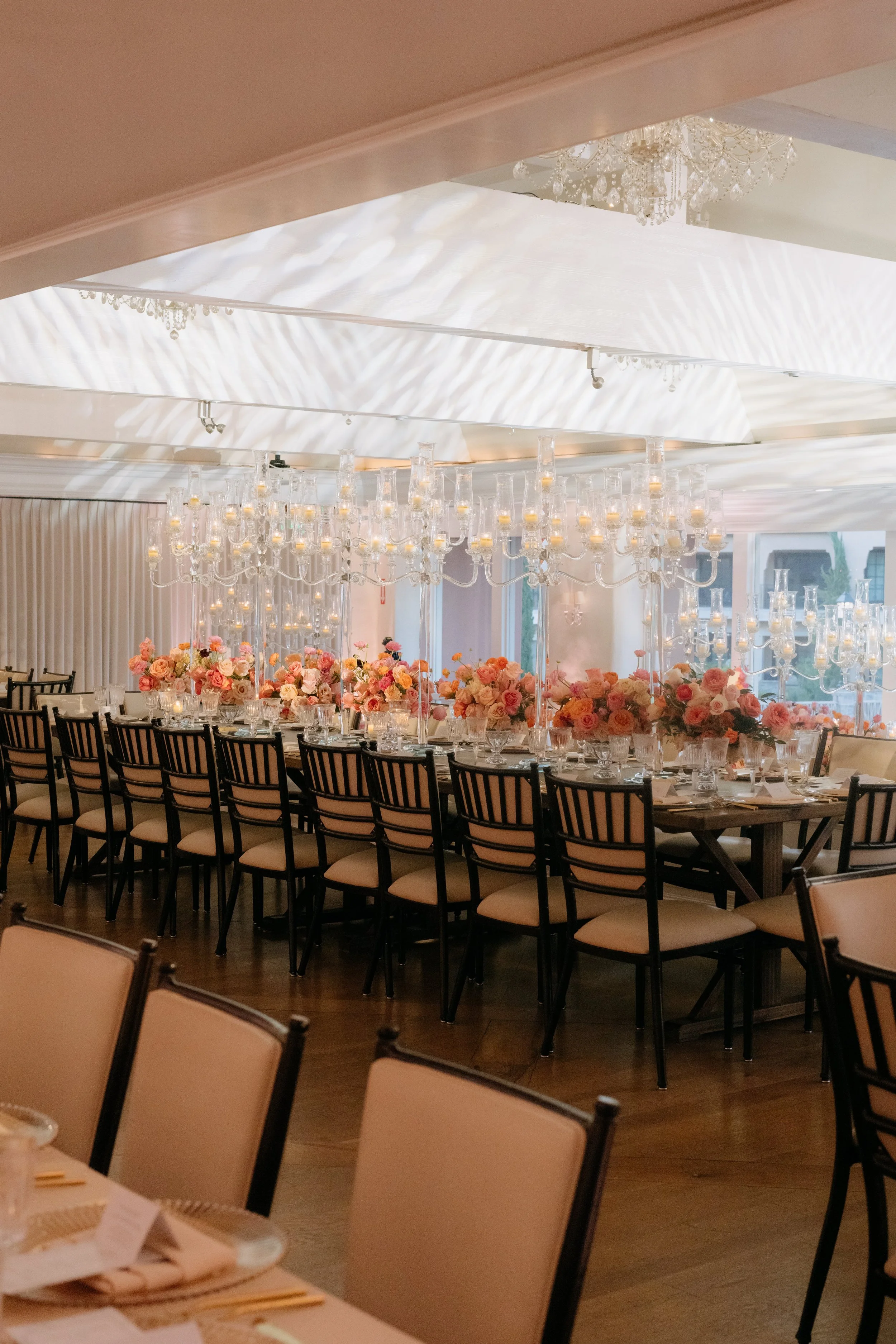 Elegant banquet hall decorated for a formal event with rows of chairs, floral centerpieces, and tall glass candle holders, illuminated by a large chandelier and patterned ceiling lighting.