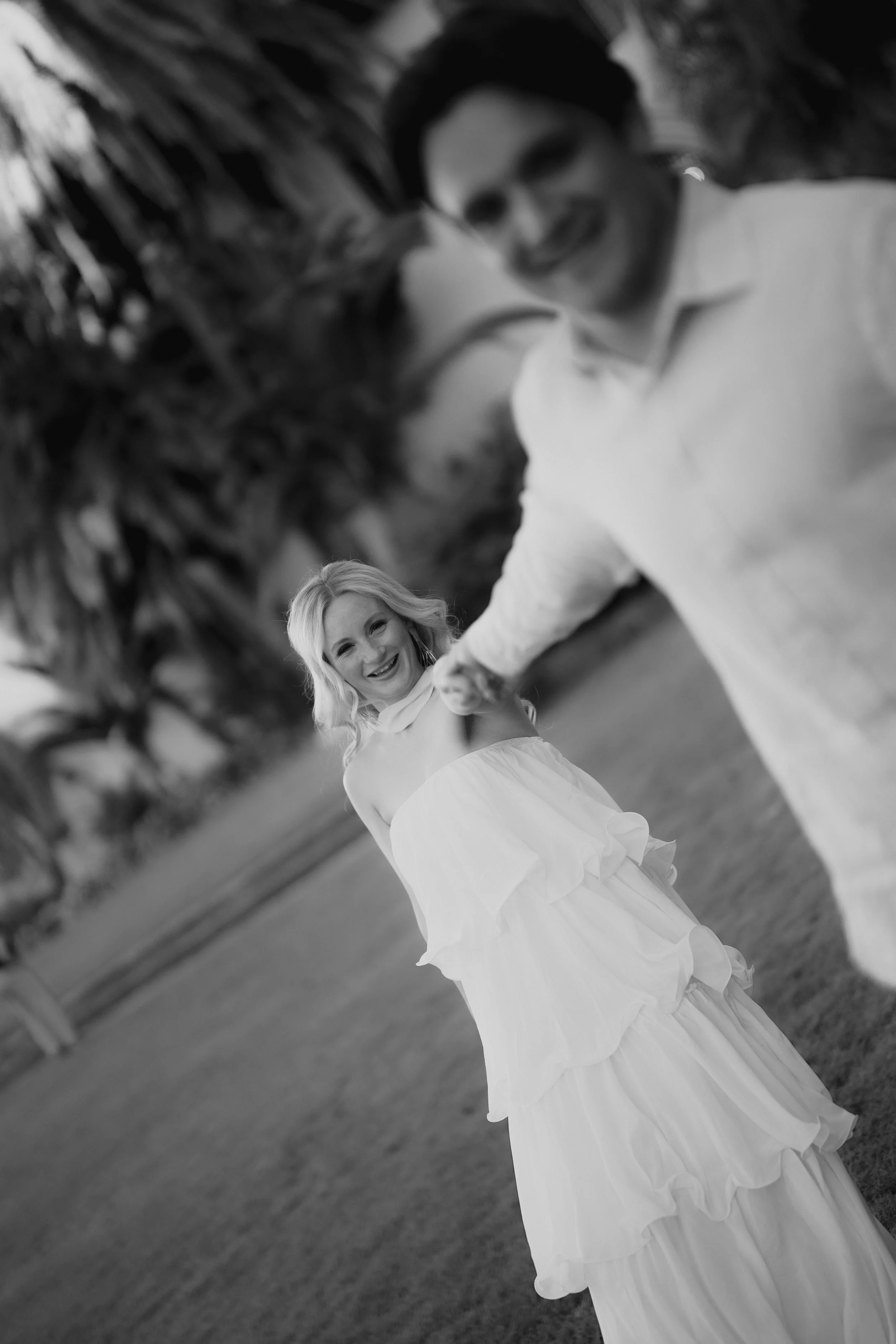 A smiling woman in a white dress holding hands with a man in a white shirt, outdoors with trees in the background.