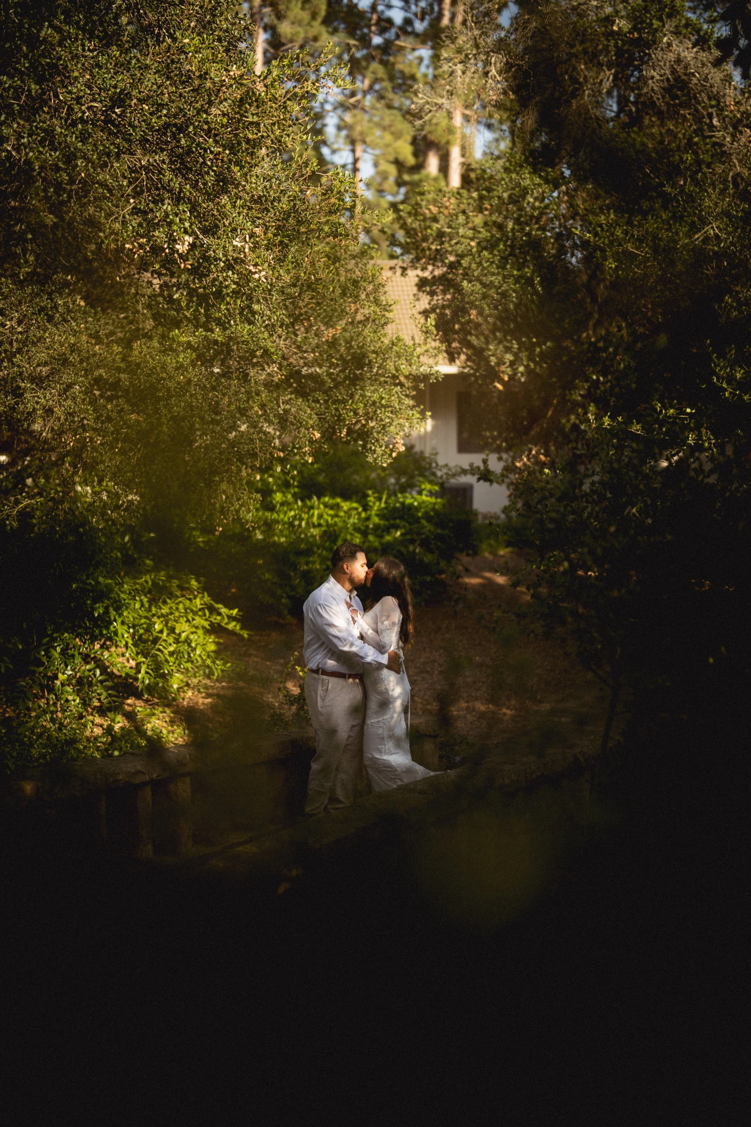 A couple dressed in white embracing and kissing outdoors in a forested area during daytime.