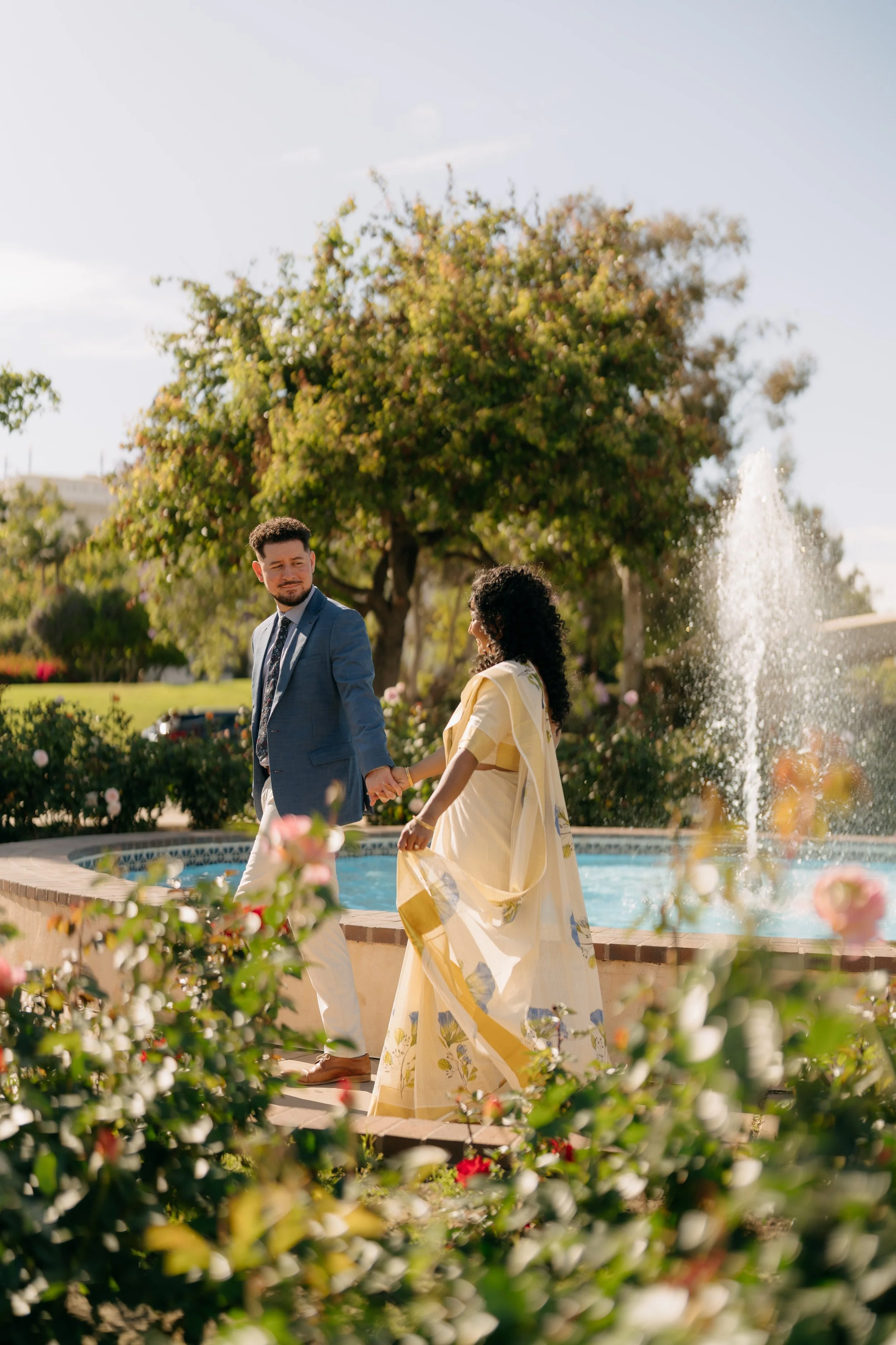 A man and a woman holding hands near a fountain in a park, with trees and flowers around them on a sunny day.