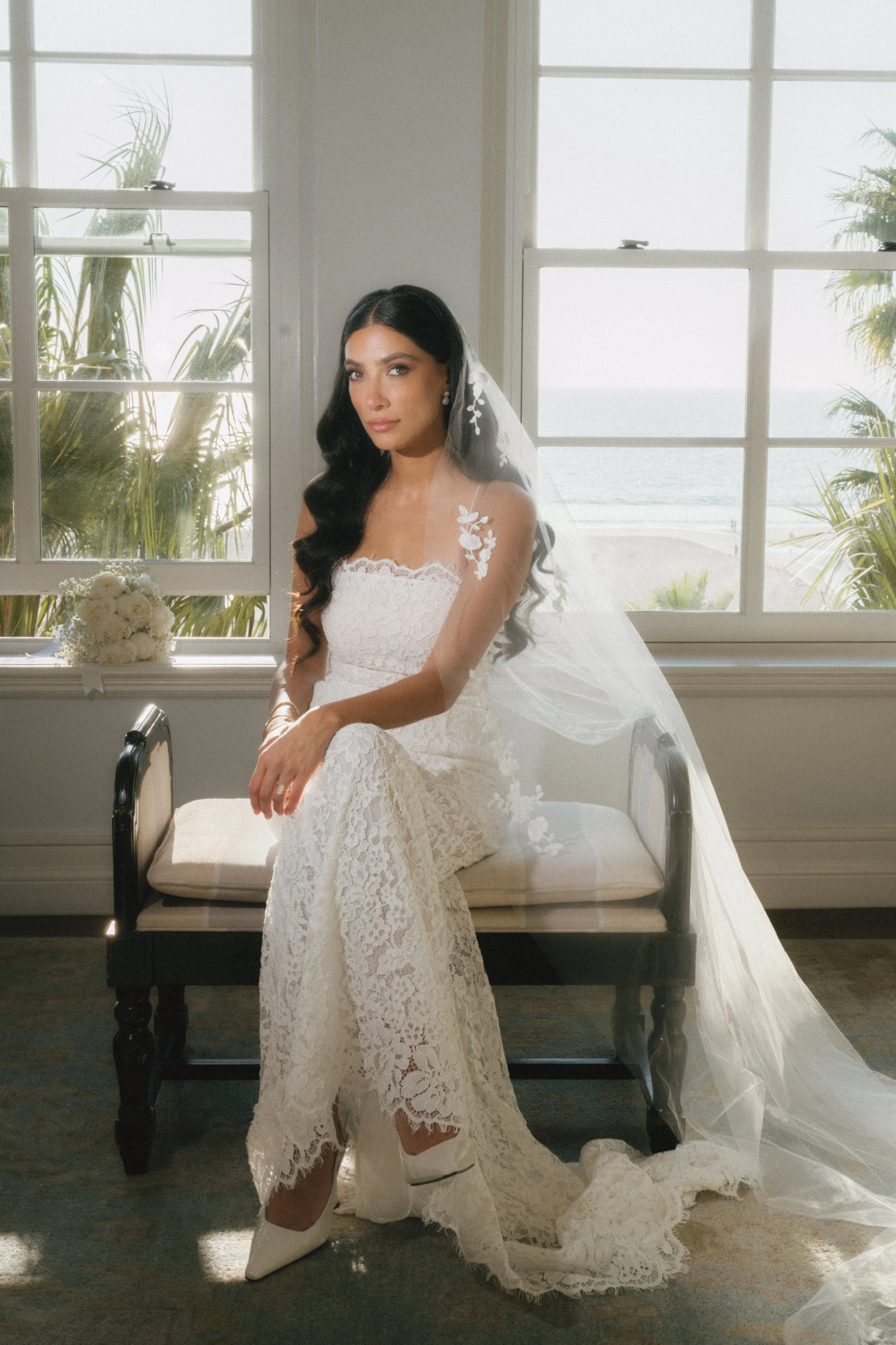 A woman in a white lace wedding dress sitting on a bench in front of large windows overlooking a beach and ocean. She has long black hair styled in waves, a veil with floral embroidery, and is wearing white high-heeled boots.