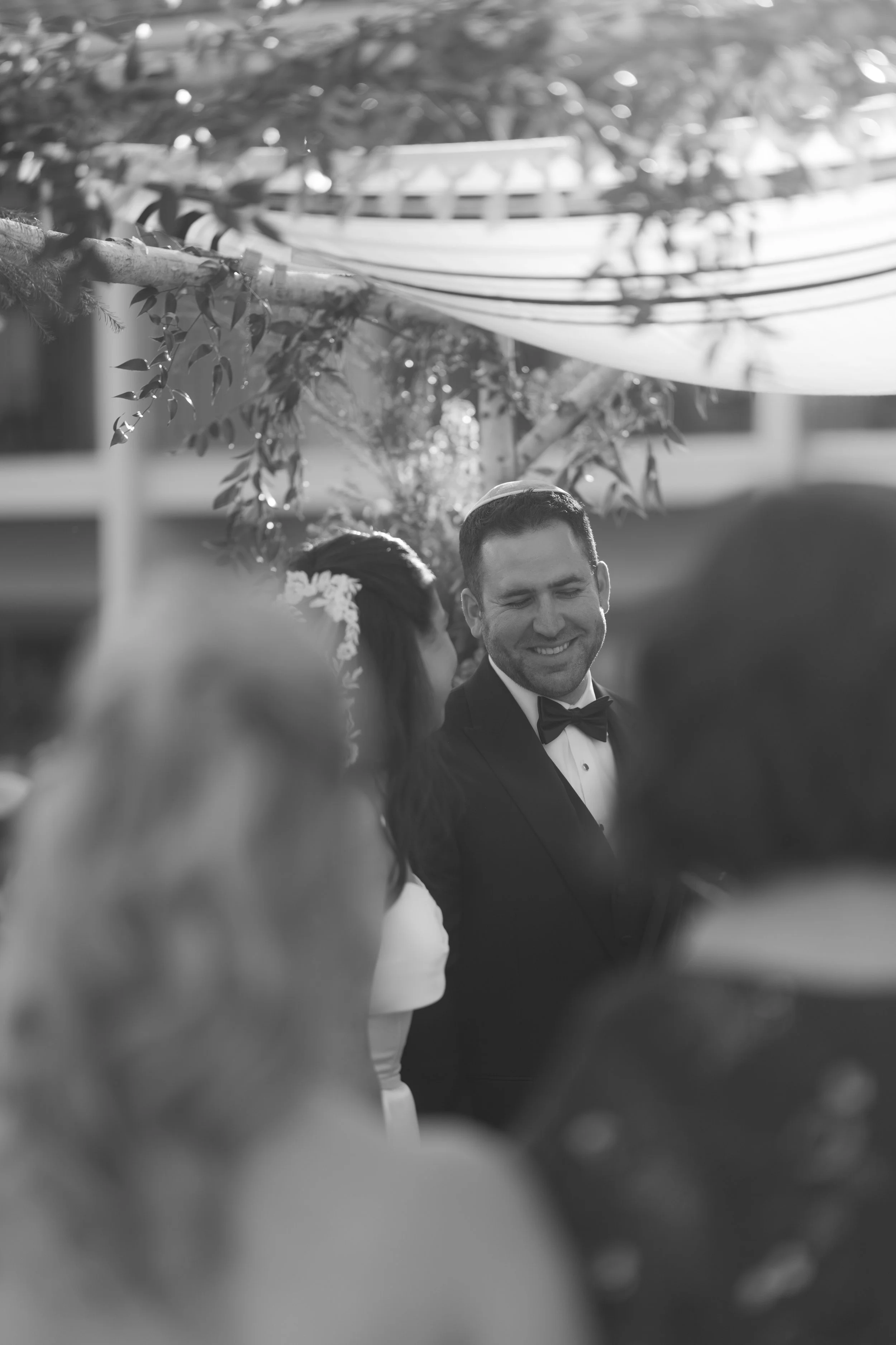 Black and white photo of a man in tuxedo smiling at a wedding ceremony, with a woman in a wedding dress and flowers on her hair visible in the foreground, and decorative hanging fabric and greenery in the background.