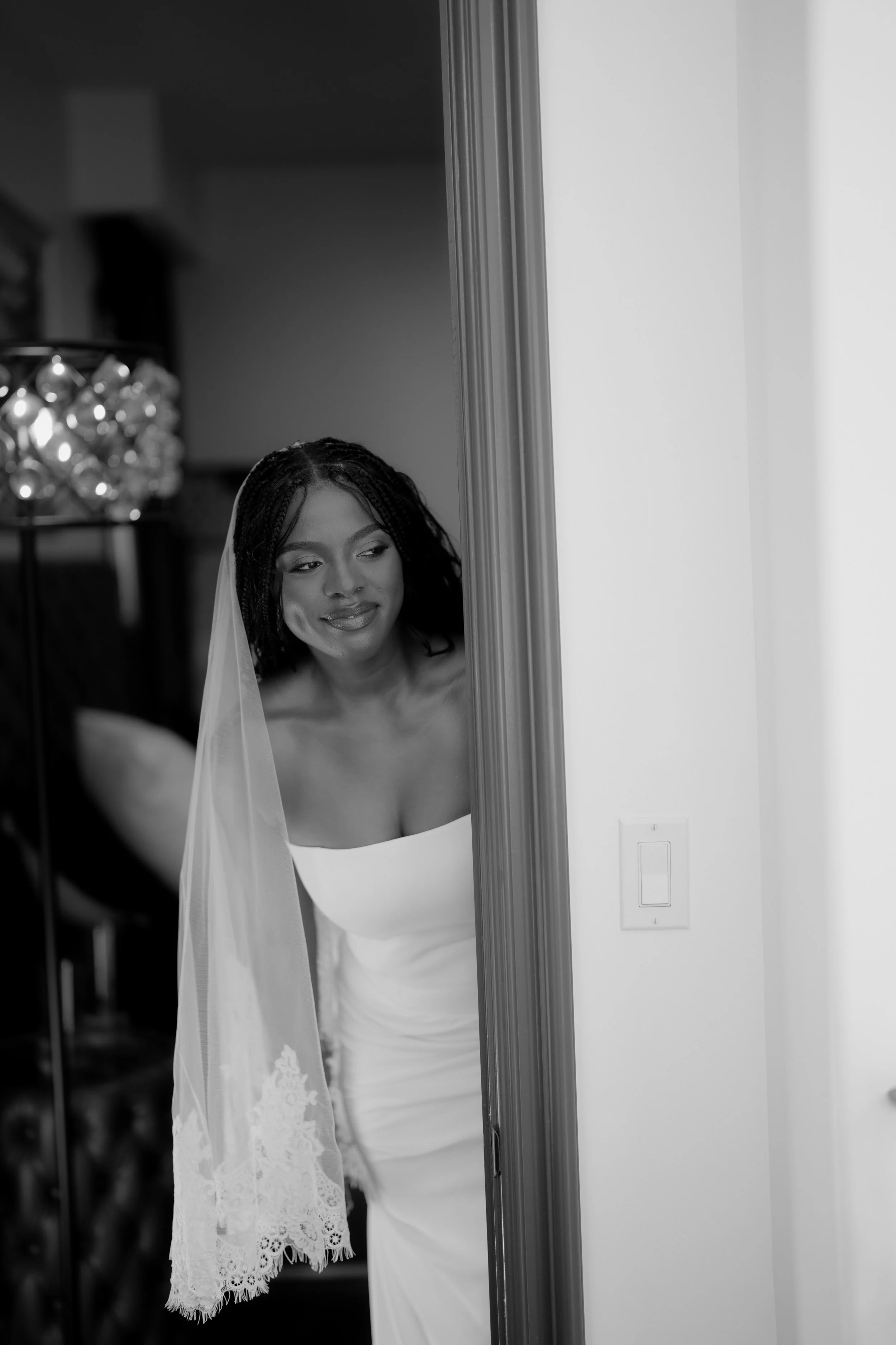 A woman wearing a white dress and veil looking at herself in a mirror, smiling, with a lace detail on her veil, in a room with a chandelier in the background.