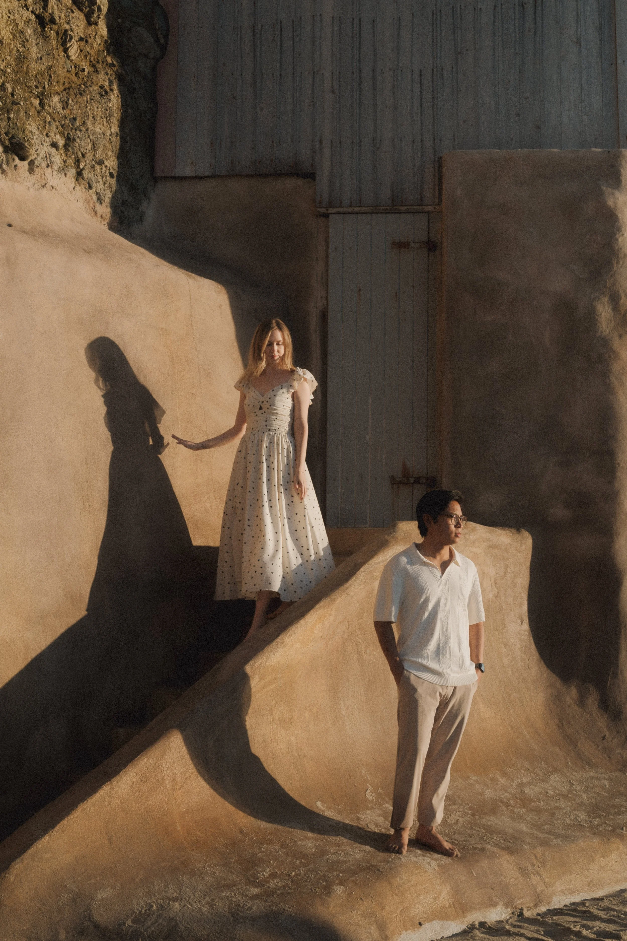 Three people standing on a rocky, sand-colored surface against a textured wall with shadows cast by sunlight.
