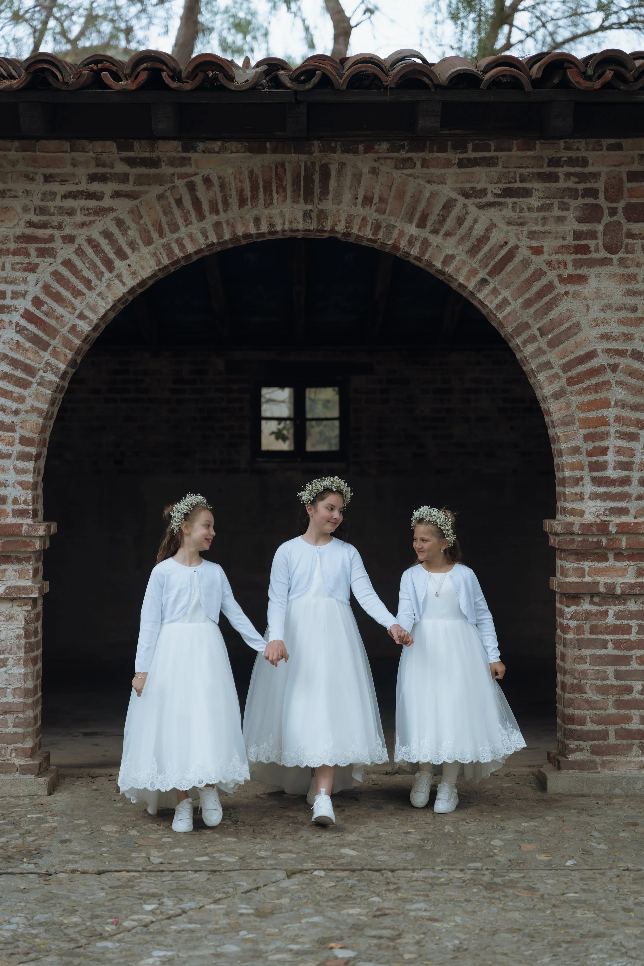 Four girls in white dresses and floral crowns holding hands, standing under a brick archway outside.