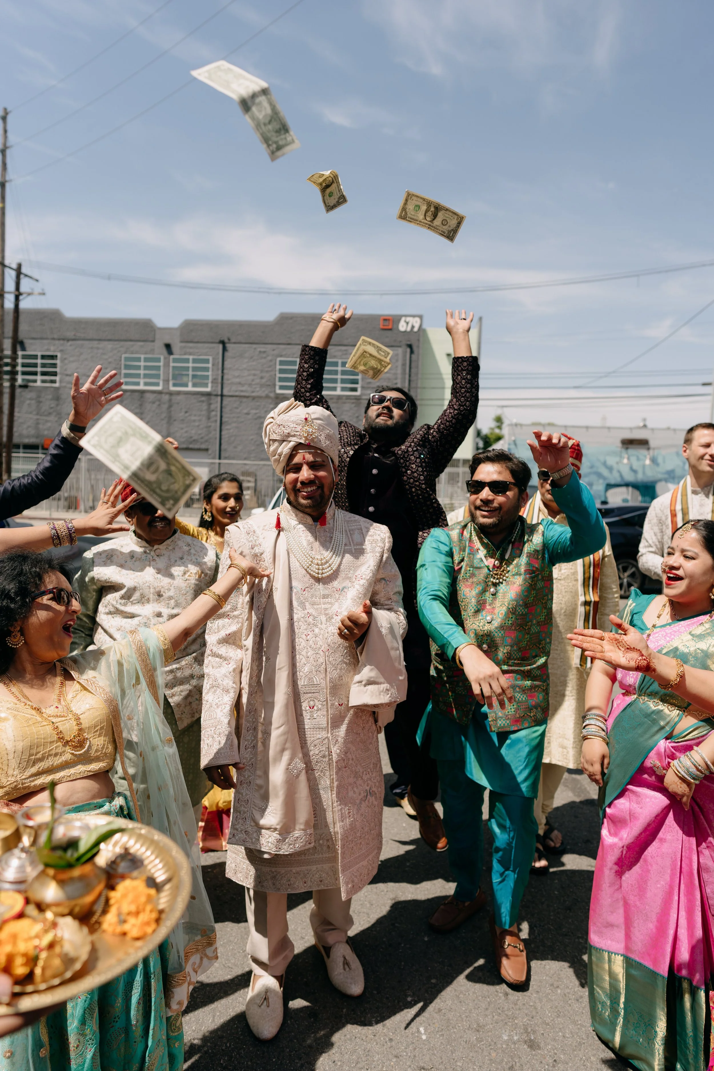 A group of people celebrating an Indian wedding outdoors, with some wearing traditional attire and throwing money into the air.