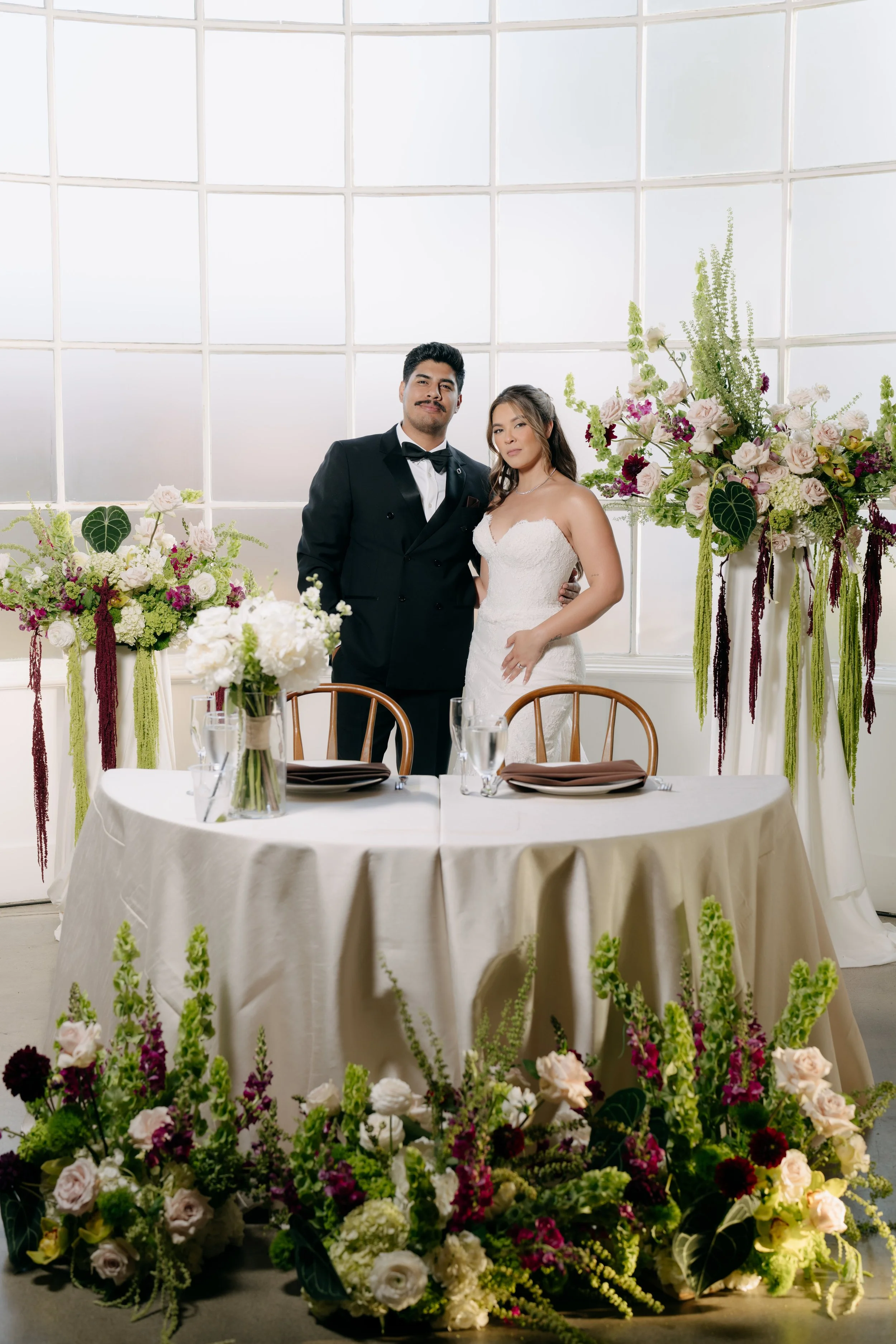 A bride and groom standing behind a table decorated with white and purple flowers, inside a bright room with large windows.