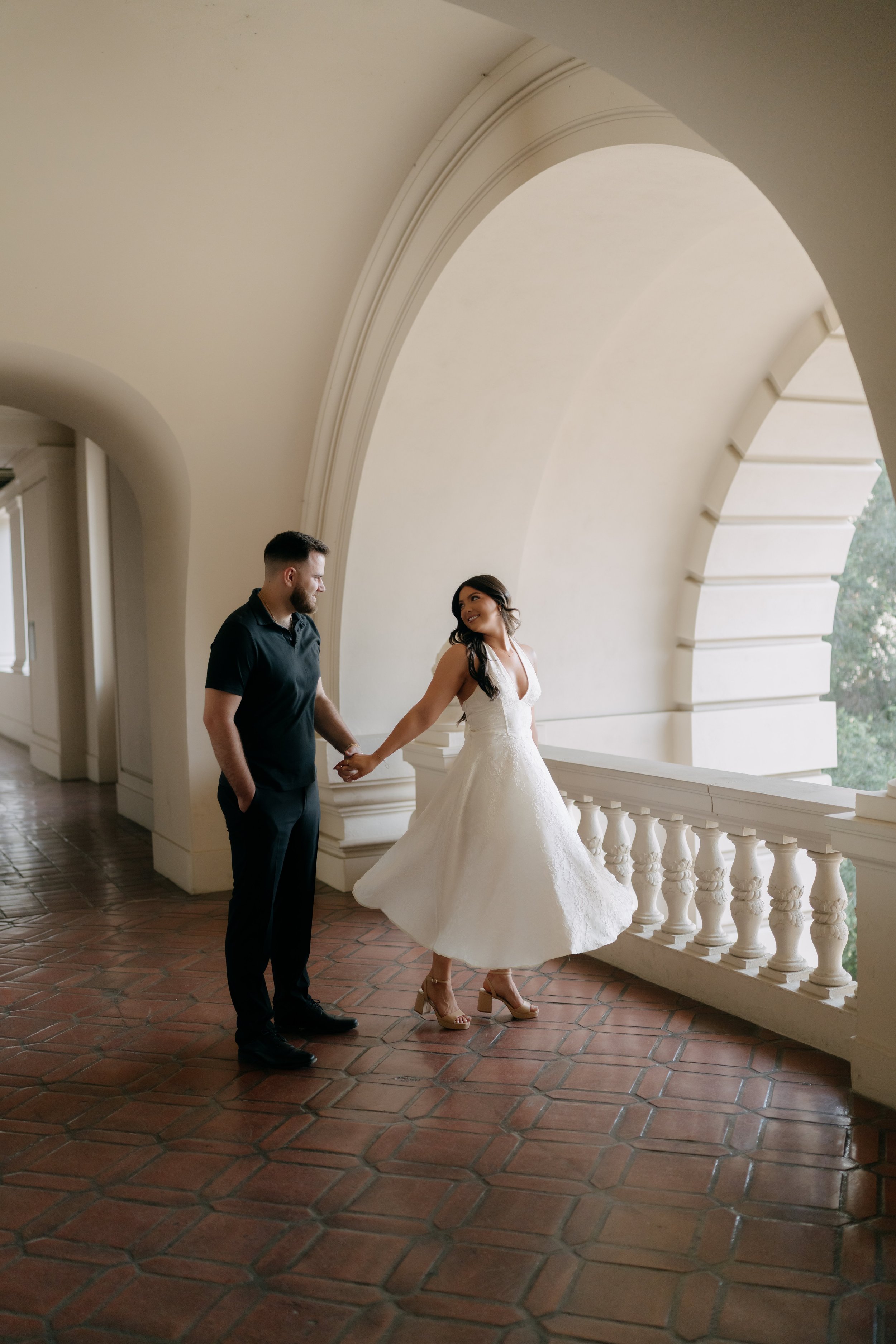 A man and a woman holding hands, standing on a balcony with an arched ceiling. The woman is wearing a white dress and high heels, and the man is dressed in dark clothing.