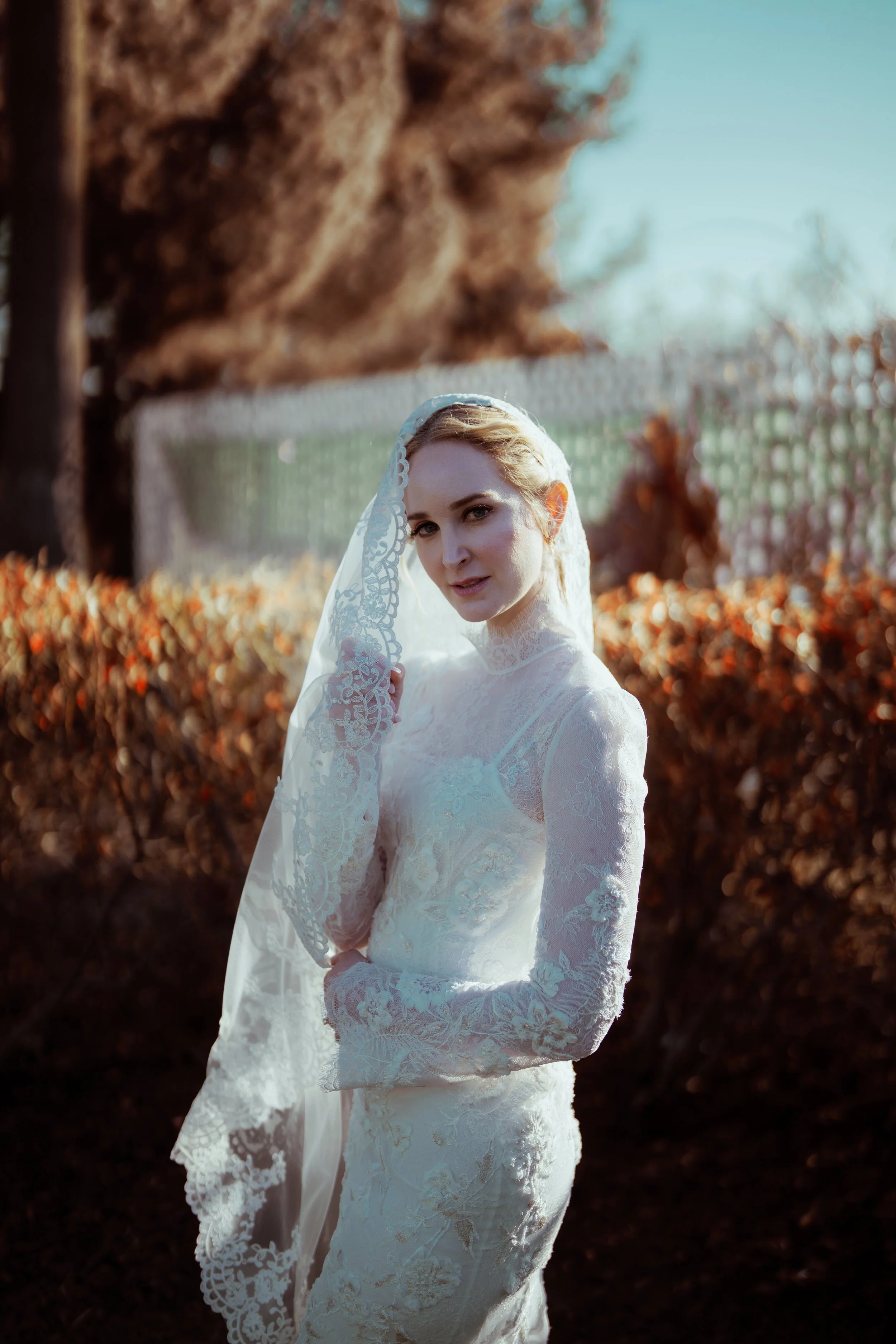 A woman in a white lace wedding dress with a lace veil outdoors during the daytime, with autumn leaves in the background.