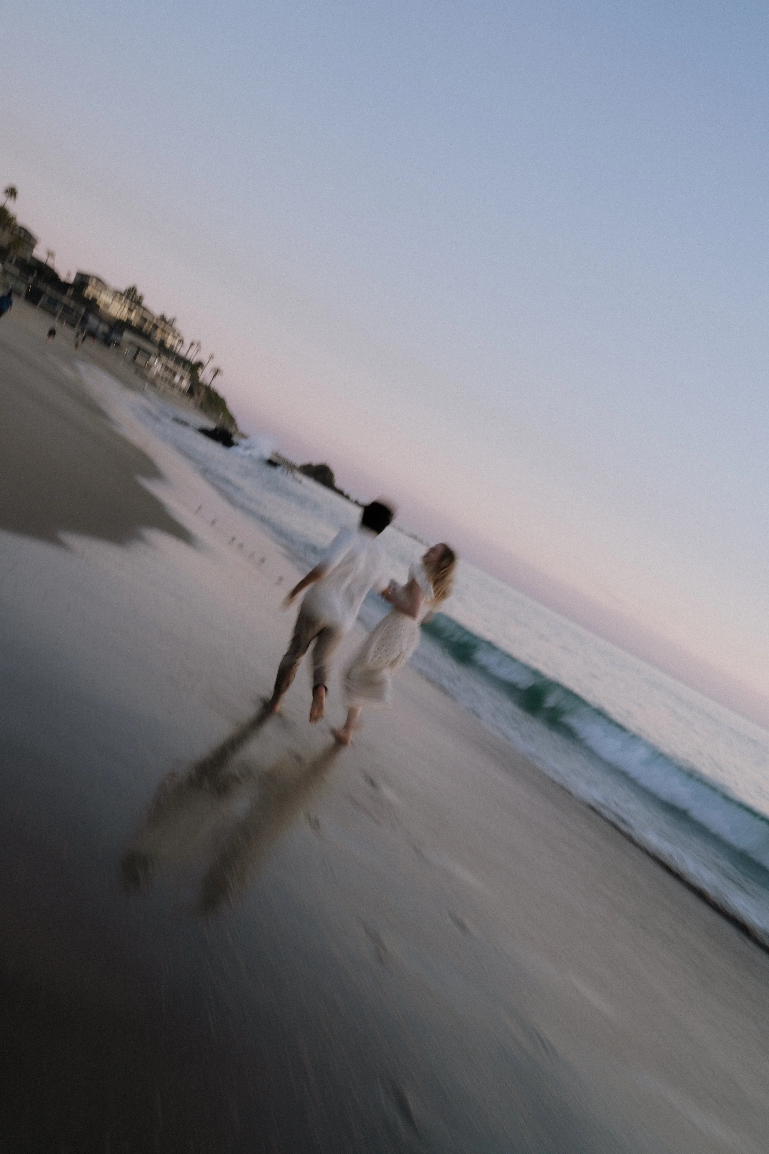 Two women walk along the beach with the ocean waves behind them during sunset, with buildings in the distance and a clear sky.