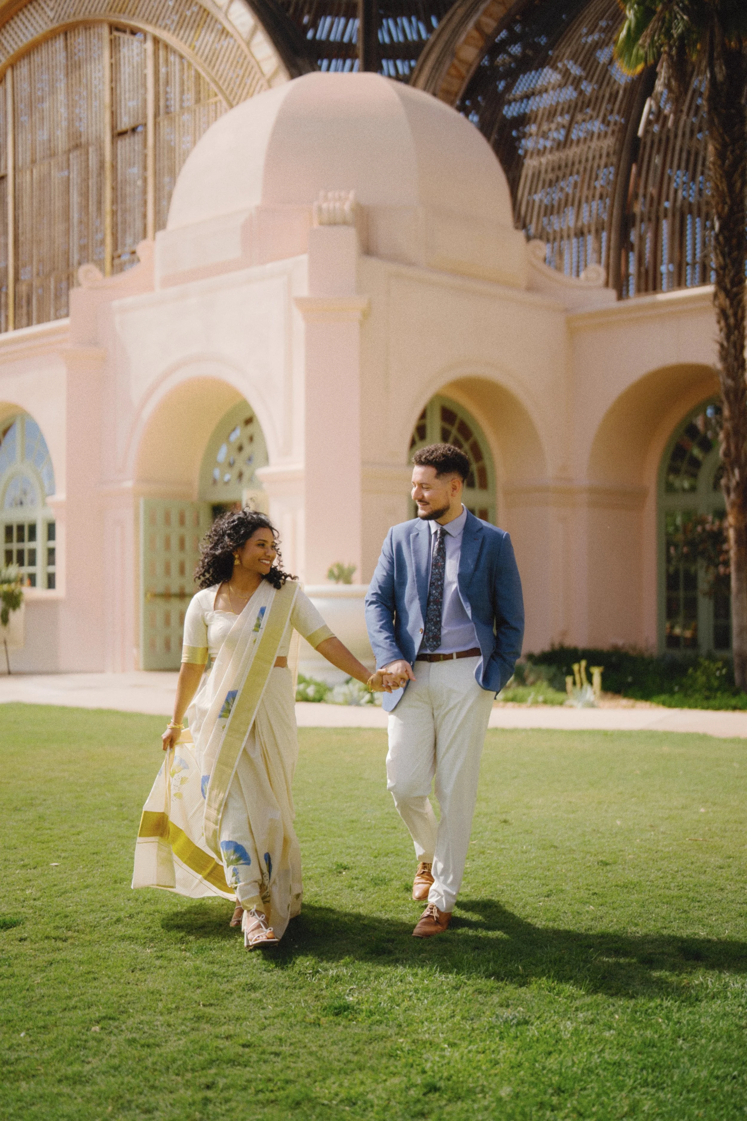 A couple walking hand in hand on a well-maintained lawn in front of a pink building with large arched windows, with a tree on the side, during daytime.