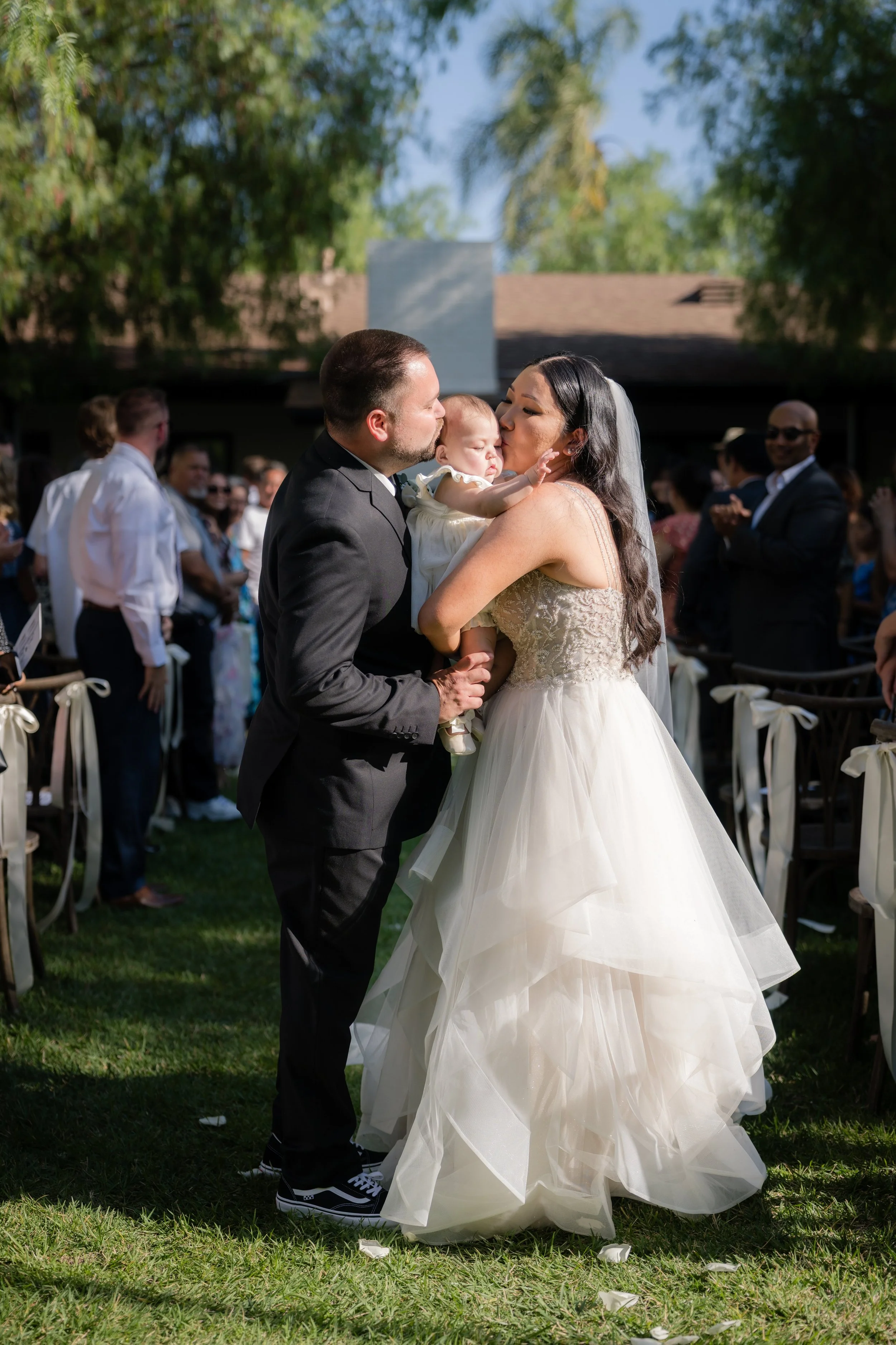A wedding ceremony outdoors with a bride, groom, and a small child, possibly their child, in a white dress. The bride and groom kiss while holding the child who is reaching out. Guests are visible in the background clapping and celebrating.