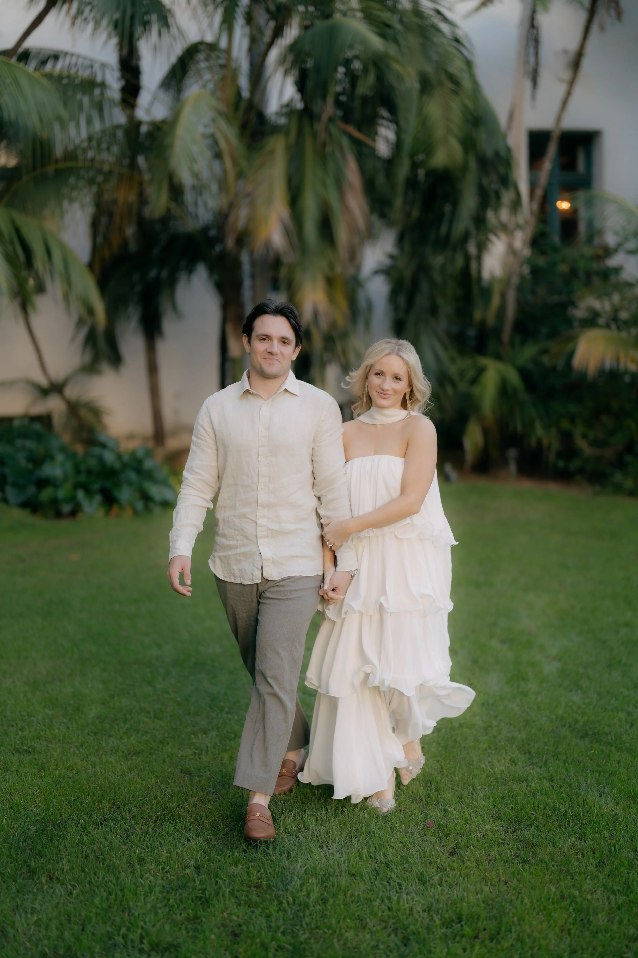 A couple walking outdoors on a grassy lawn, holding hands, with tropical plants and a white building in the background.
