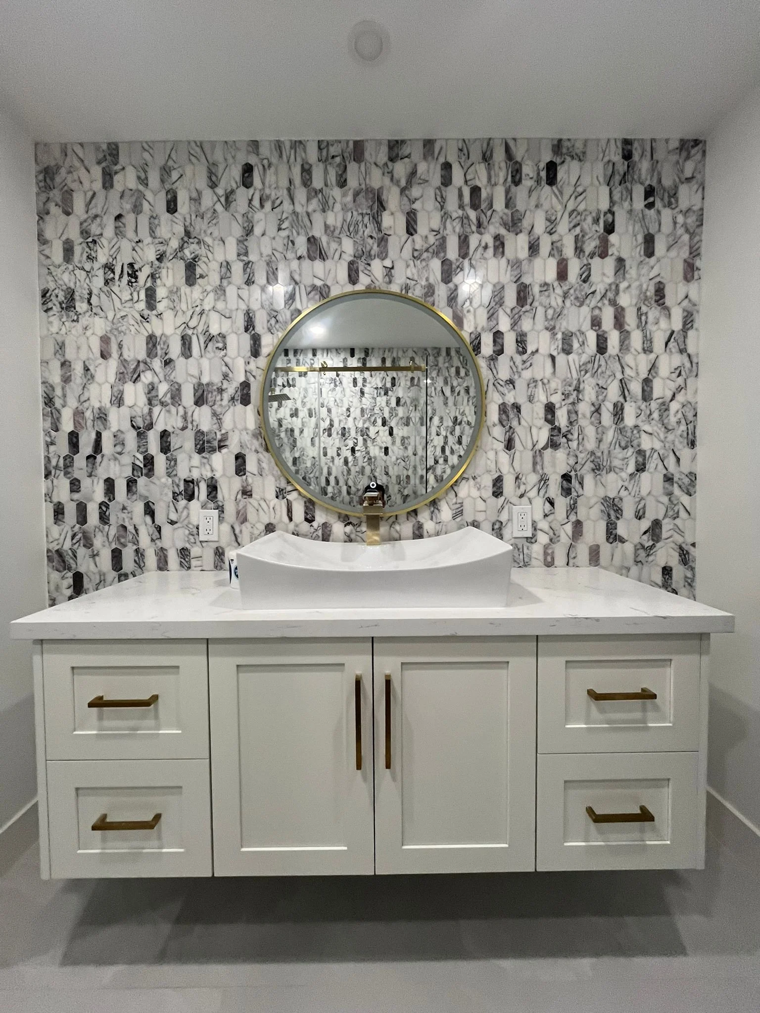 Modern bathroom vanity with white countertop, vessel sink, and round mirror. Background features a patterned tile wall. Cabinets have gold handles.