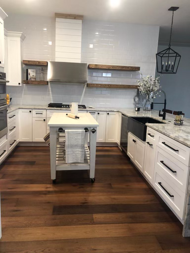 Modern kitchen interior with white cabinets, wooden floor, marble countertop, and a center island with a towel rack. Features include a farmhouse sink, stainless steel appliances, floating wooden shelves, and a hanging light fixture.