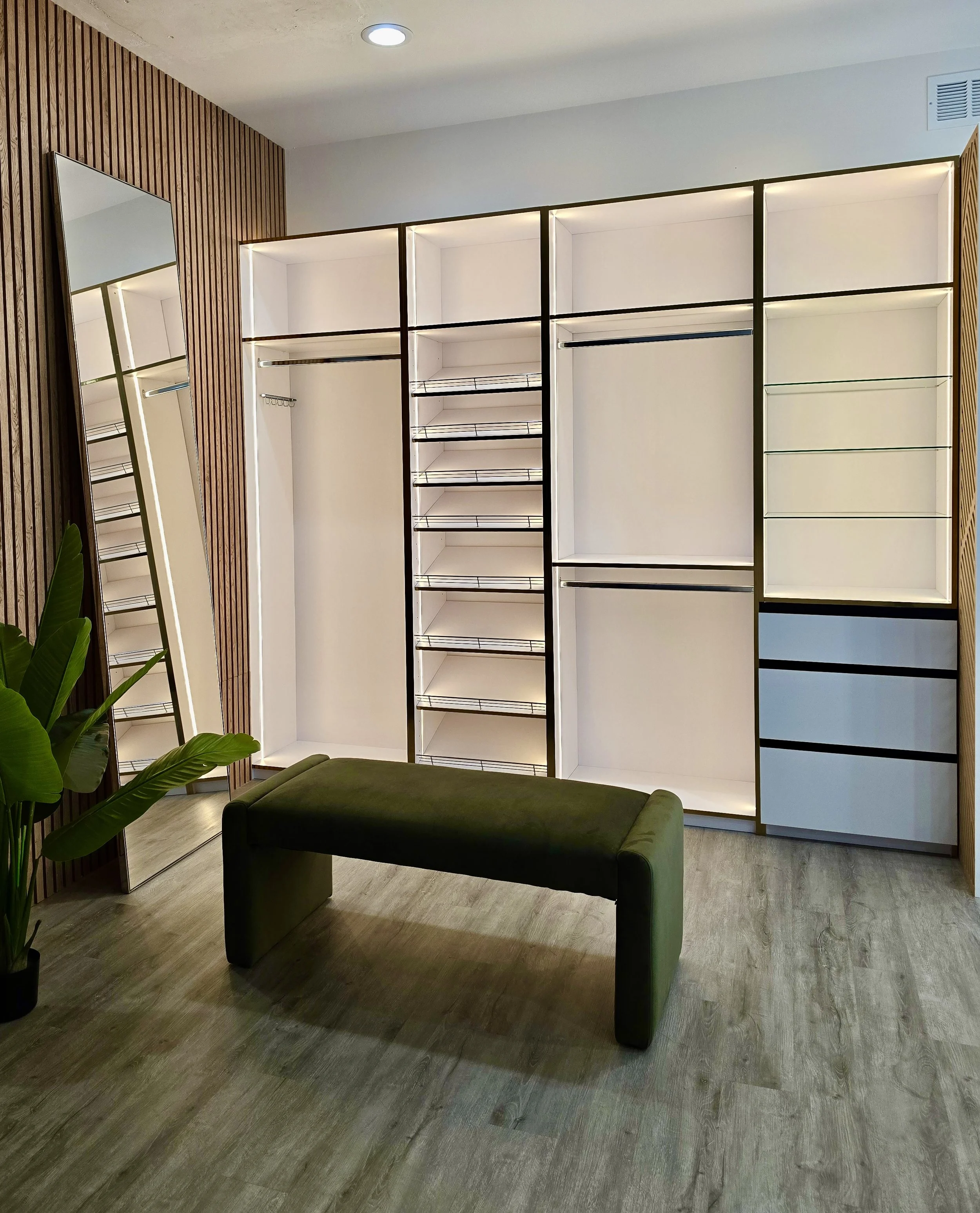 Modern walk-in closet with built-in shelves and drawers, large mirror, green bench, plant, and wood paneling on walls.